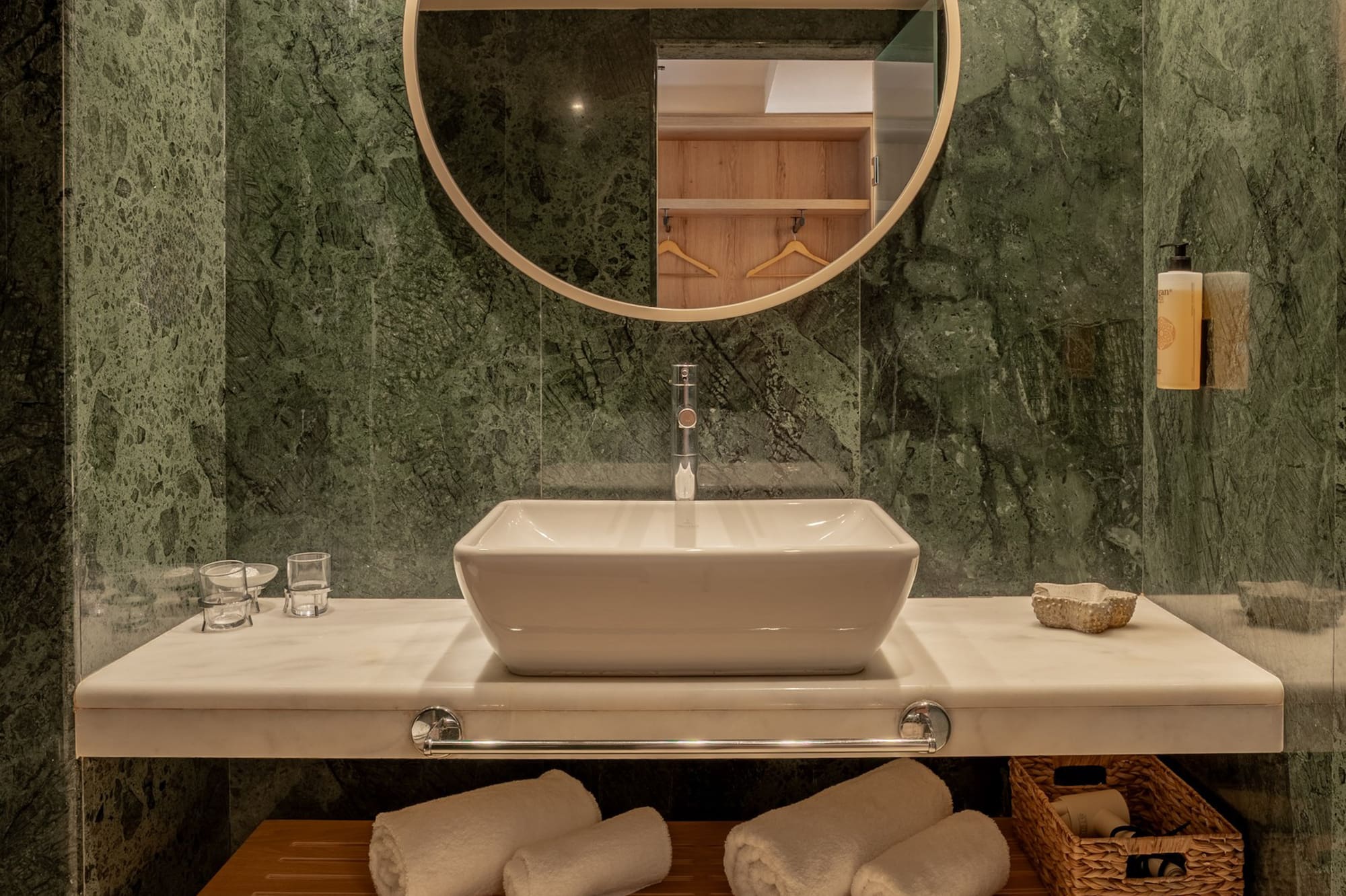 Modern bathroom with green marble, vessel sink, round mirror, and wood storage bench.
