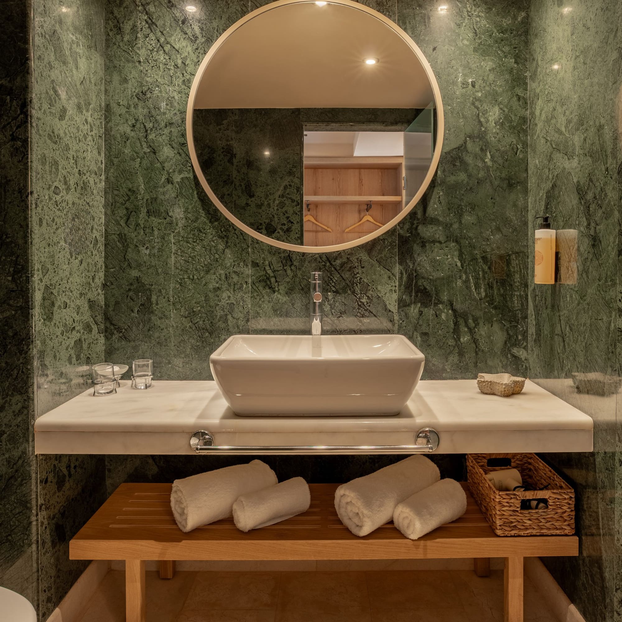Modern bathroom with green marble, vessel sink, round mirror, and wood storage bench.