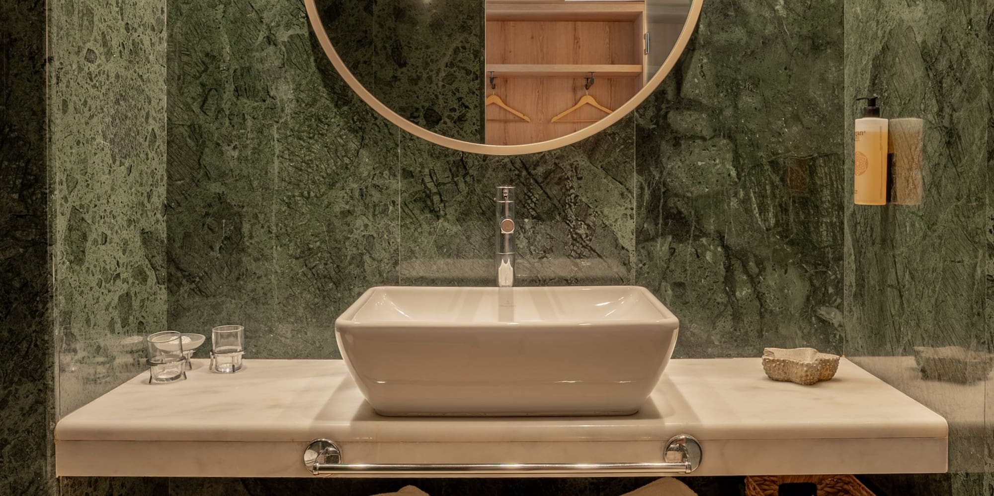 Modern bathroom with green marble, vessel sink, round mirror, and wood storage bench.