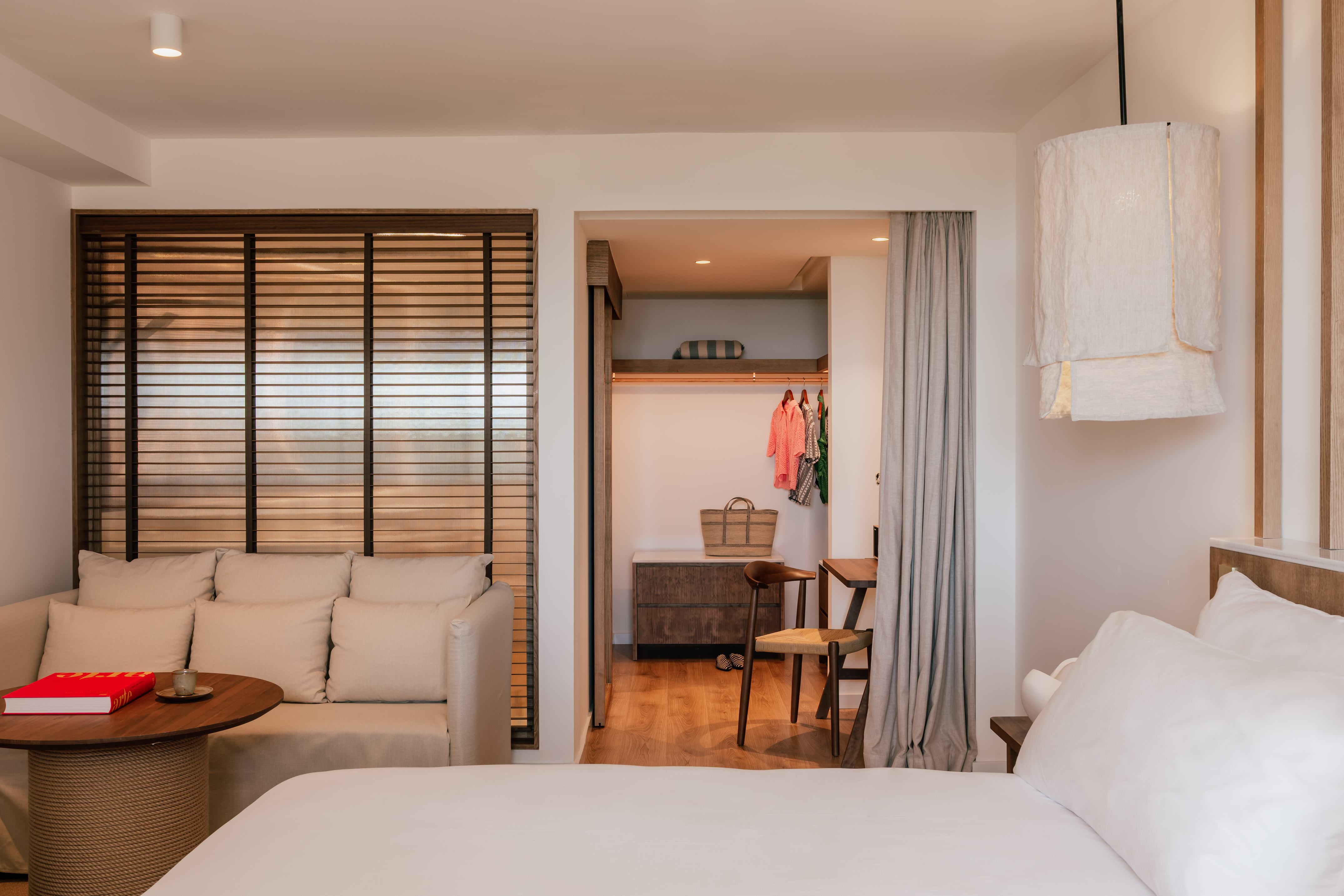 Elegant hotel bedroom with white bed, sofa, wooden blinds, and open wardrobe.