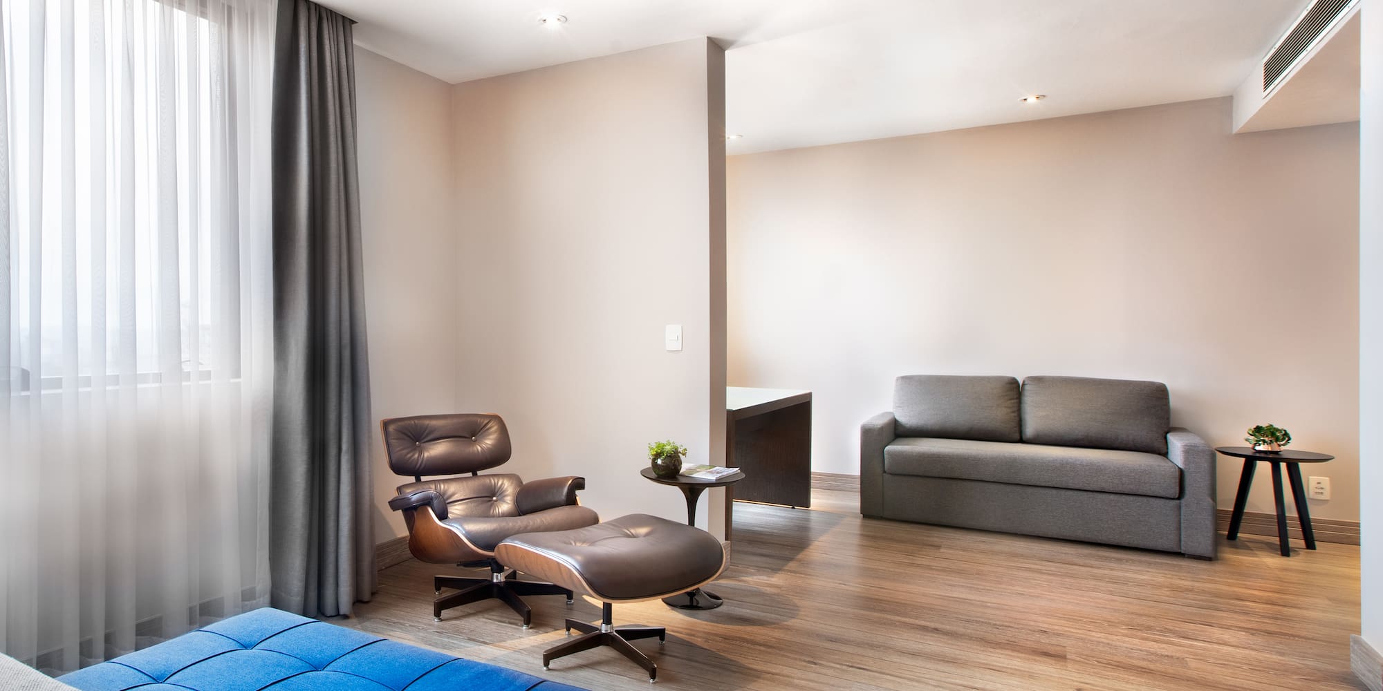 Modern hotel studio with leather lounge chair, grey sofa, and wood floor.