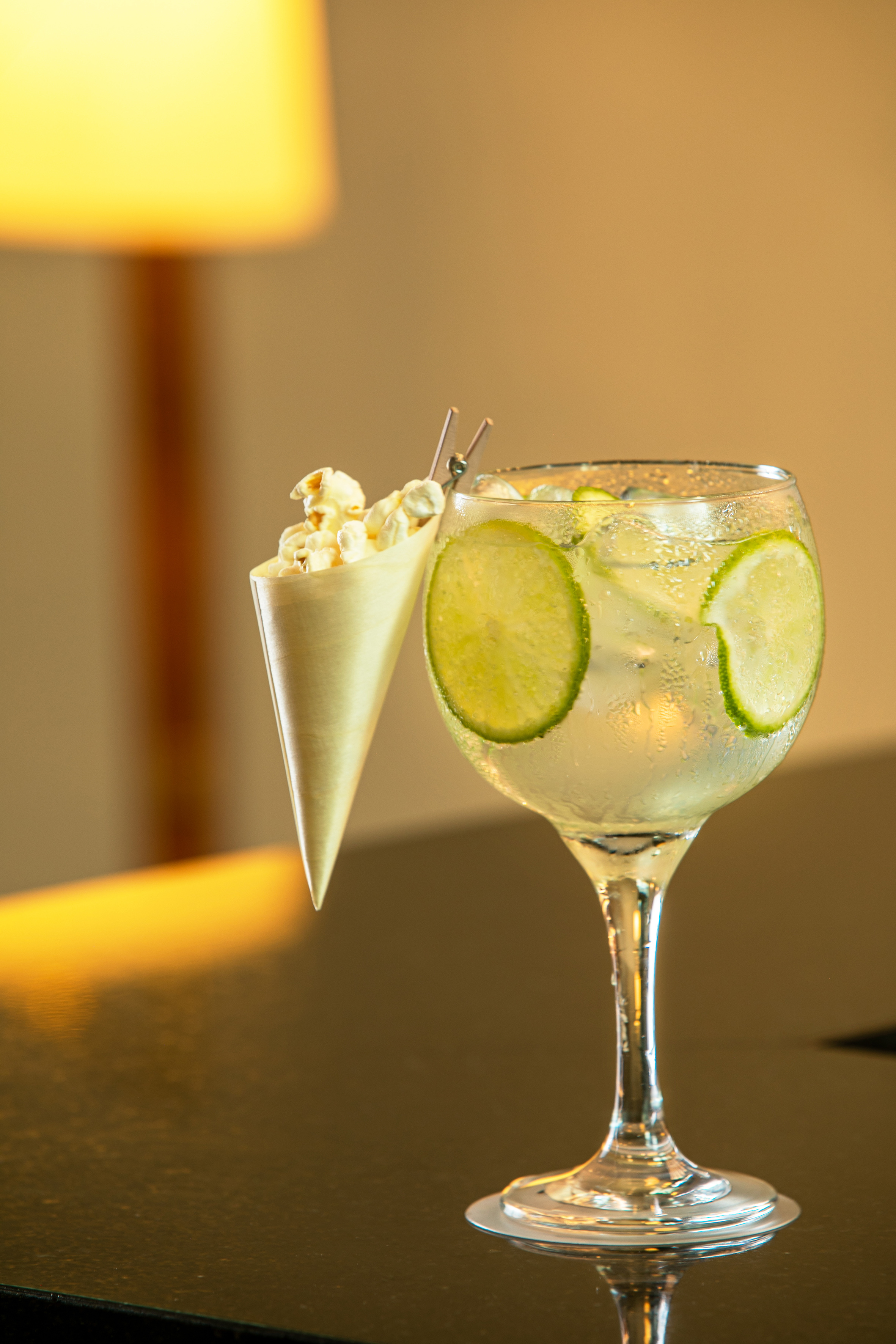Refreshing gin and tonic with lime, ice, and popcorn on a dark bar.