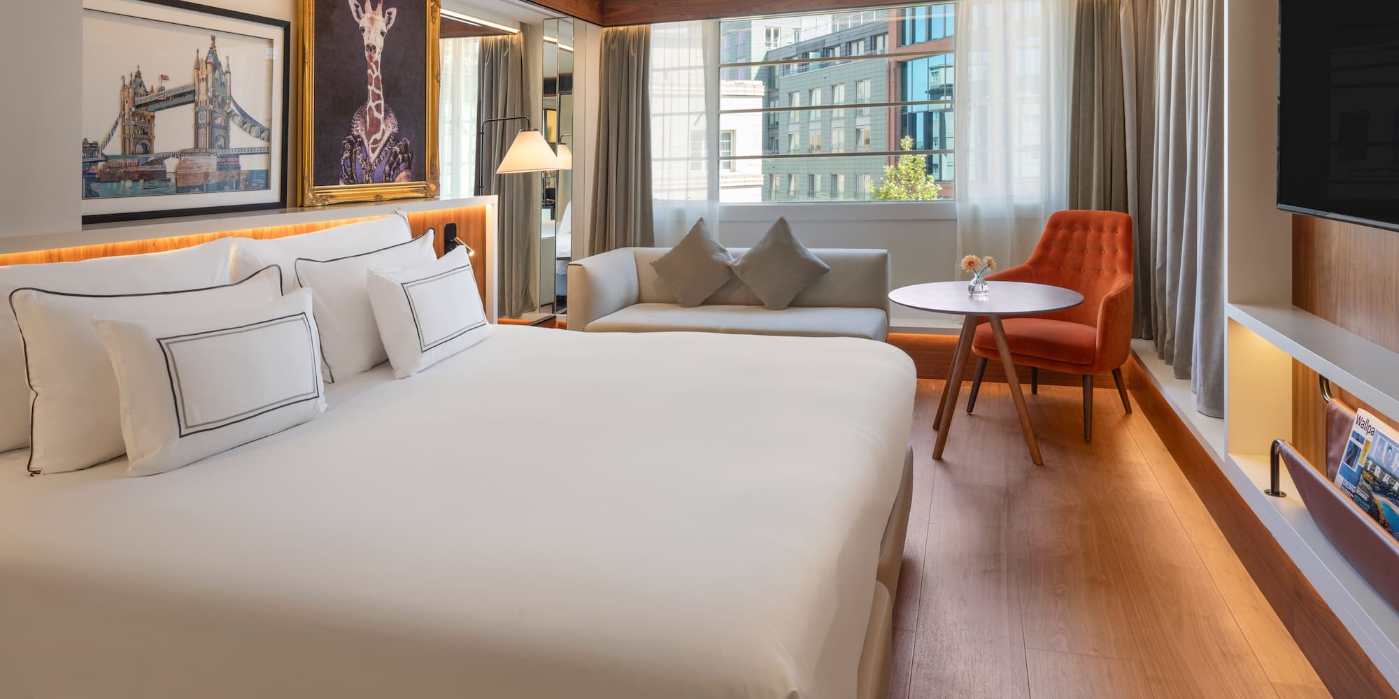 Modern hotel room with king bed, sofa, city view window, and unique art.