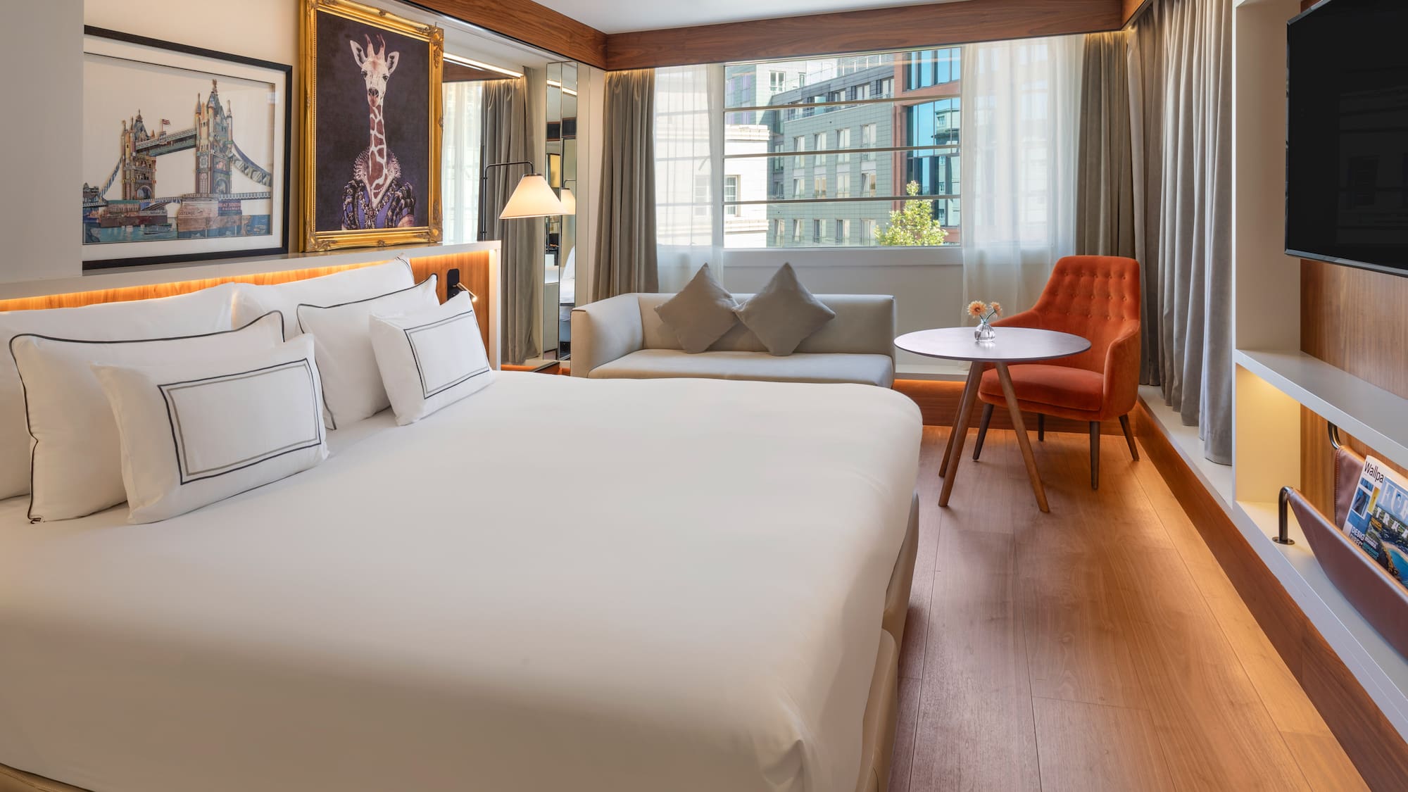 Modern hotel room with king bed, sofa, city view window, and unique art.