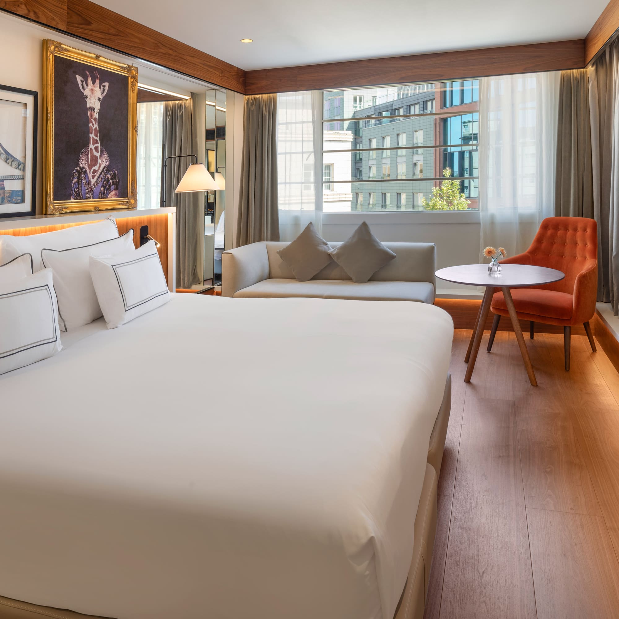 Modern hotel room with king bed, sofa, city view window, and unique art.