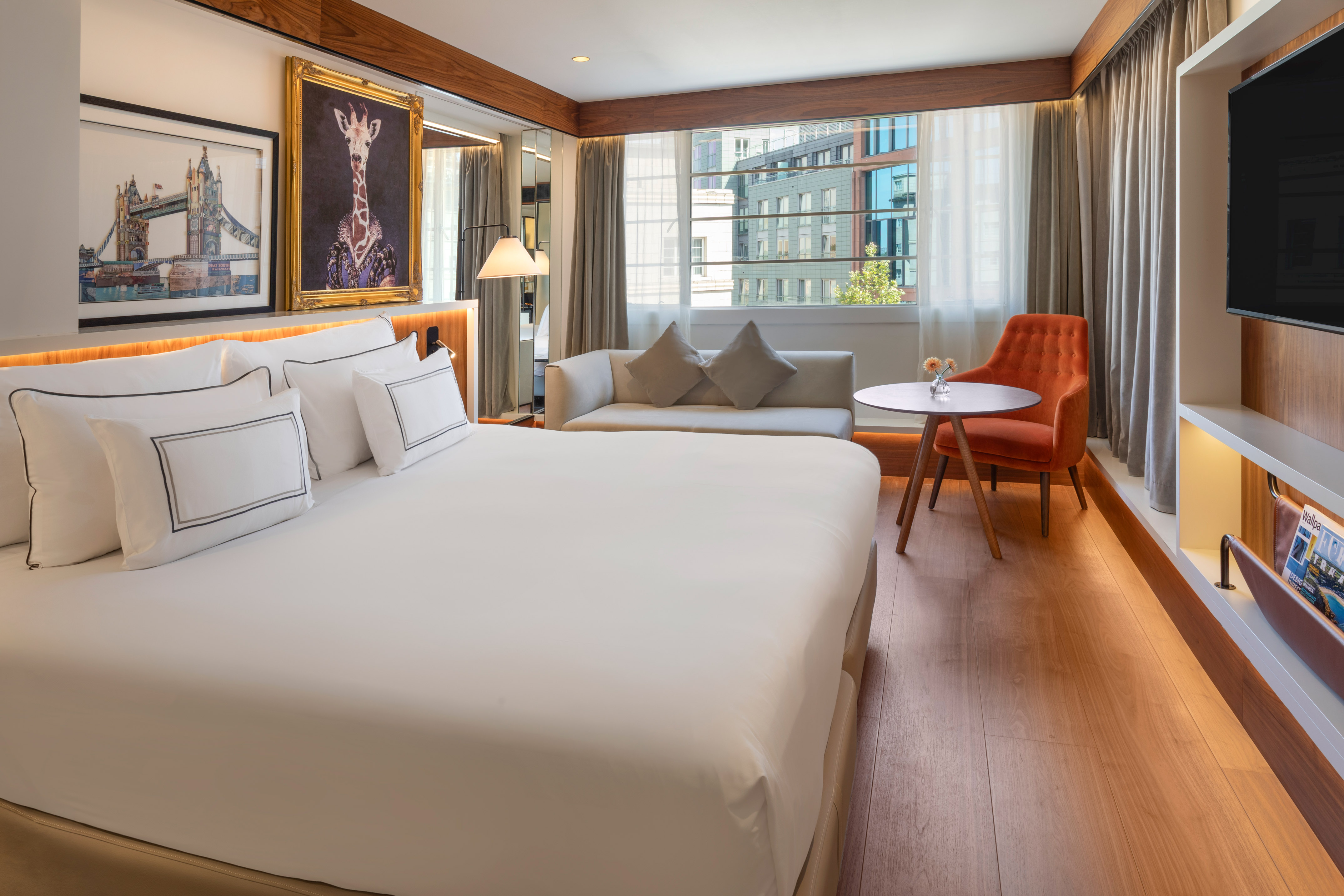 Modern hotel room with king bed, sofa, city view window, and unique art.