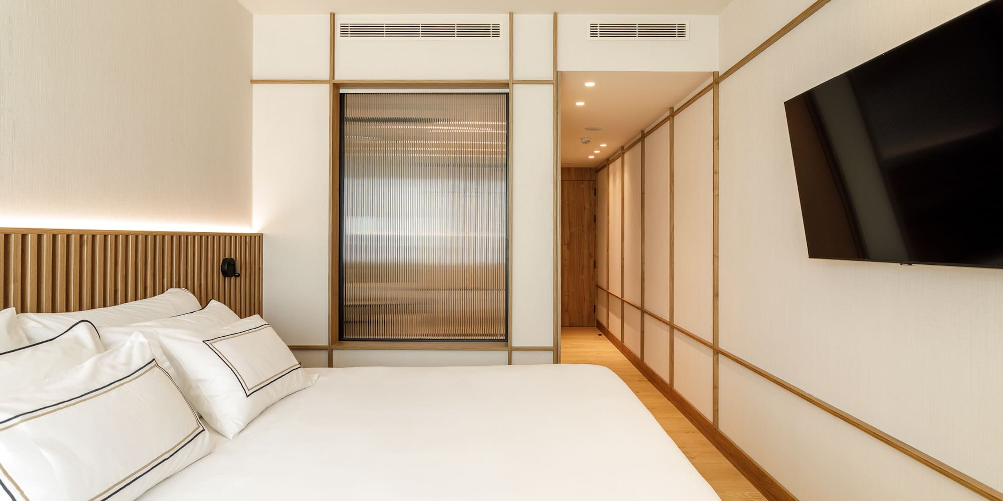 Modern hotel bedroom with white bed, wood headboard, fluted glass, and TV.