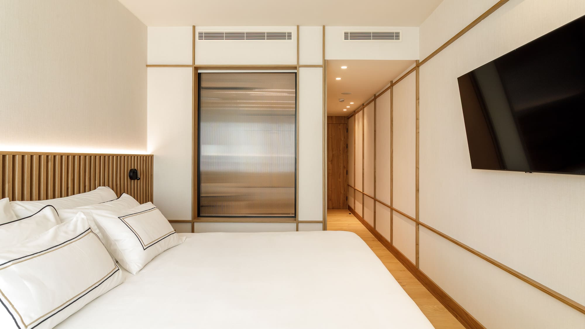 Modern hotel room with white bed, wooden headboard, and flat-screen TV.