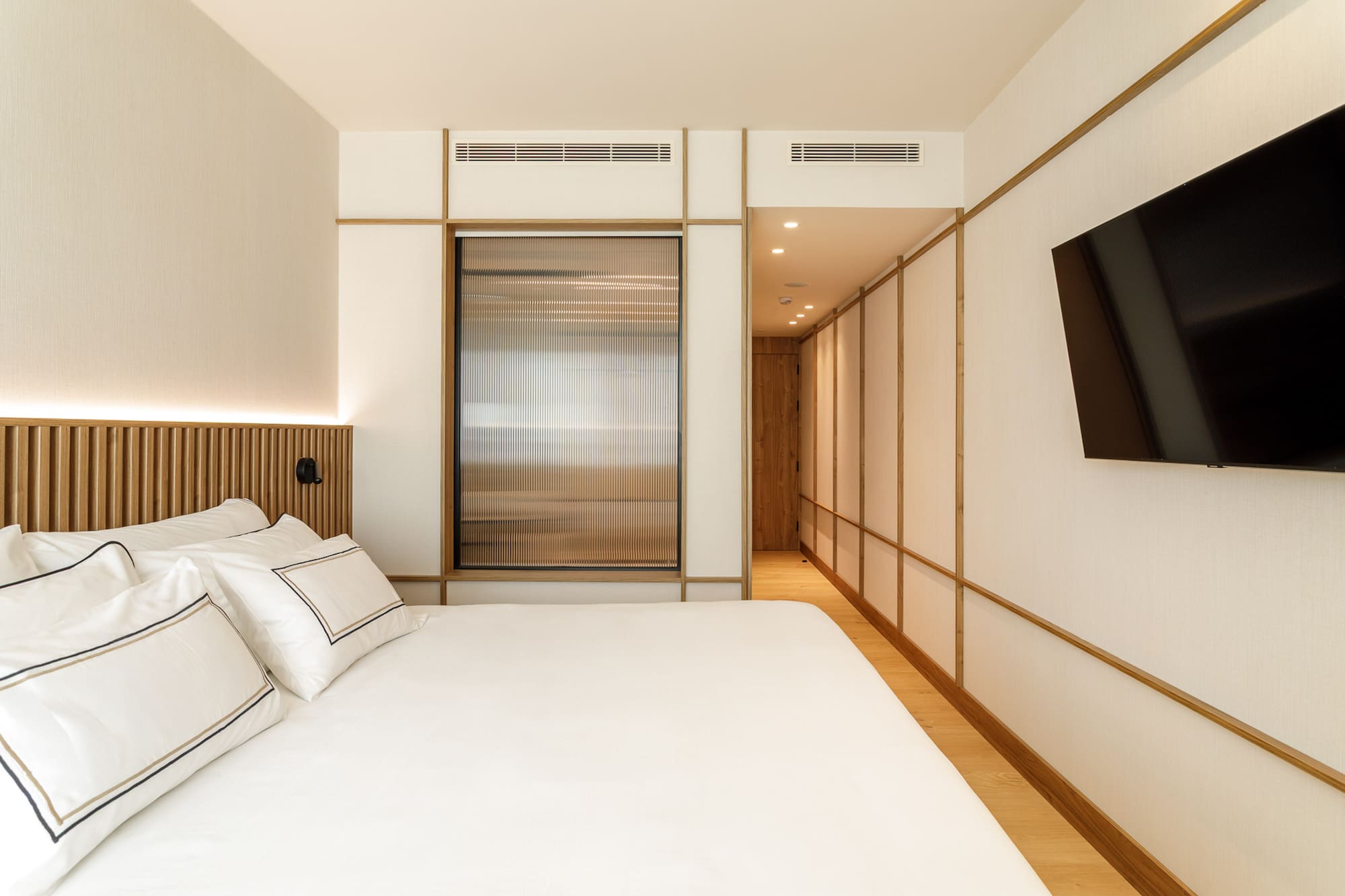 Modern minimalist bedroom with white bed, wooden accents, and wall-mounted TV.