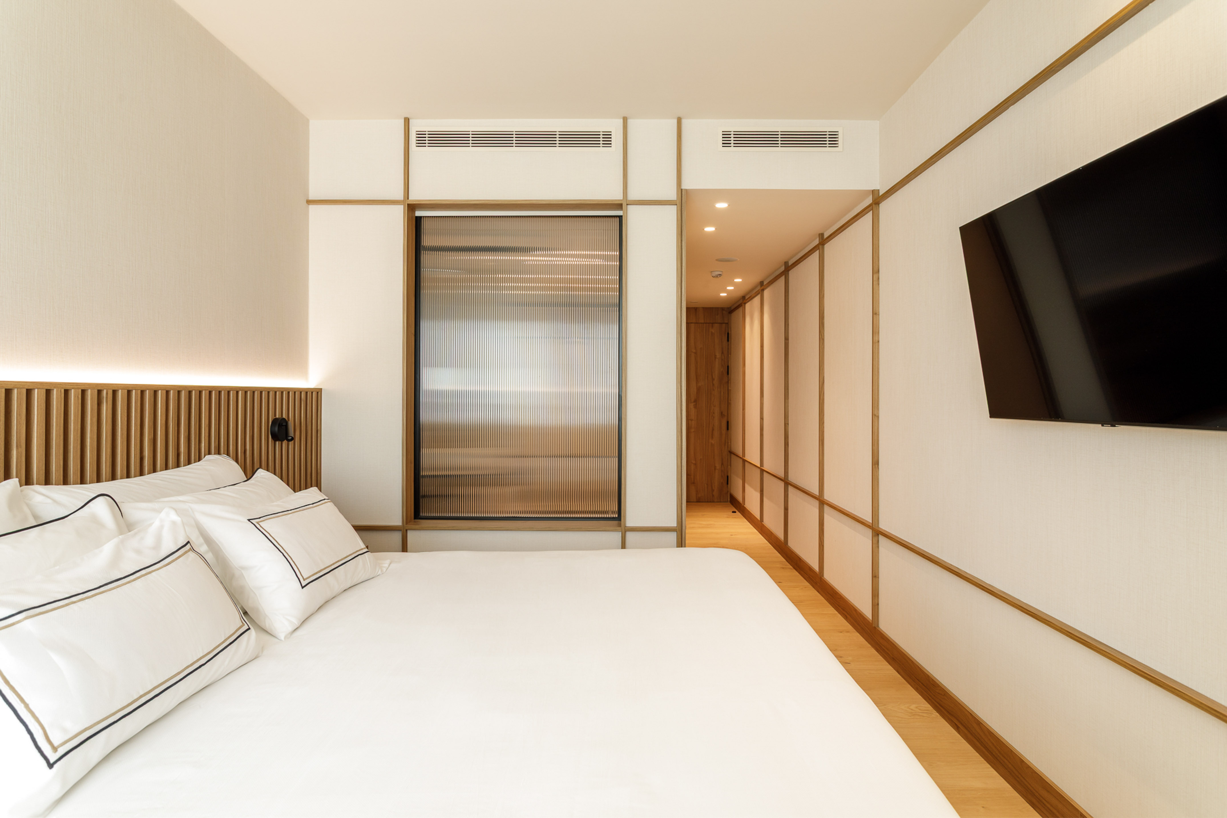 Modern hotel room with white bed, wooden headboard, and flat-screen TV.