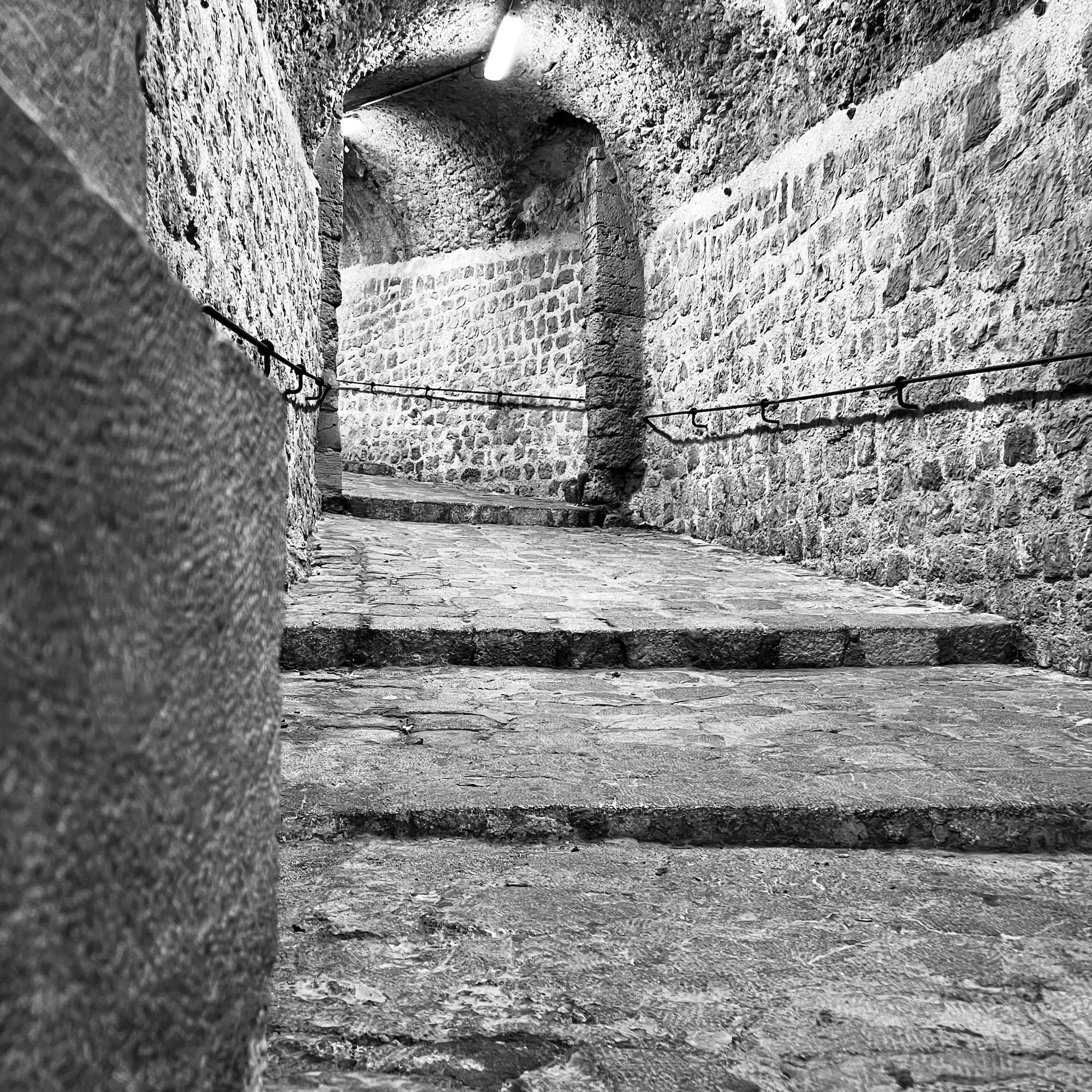 Black and white stone arched tunnel with illuminated steps and handrail.