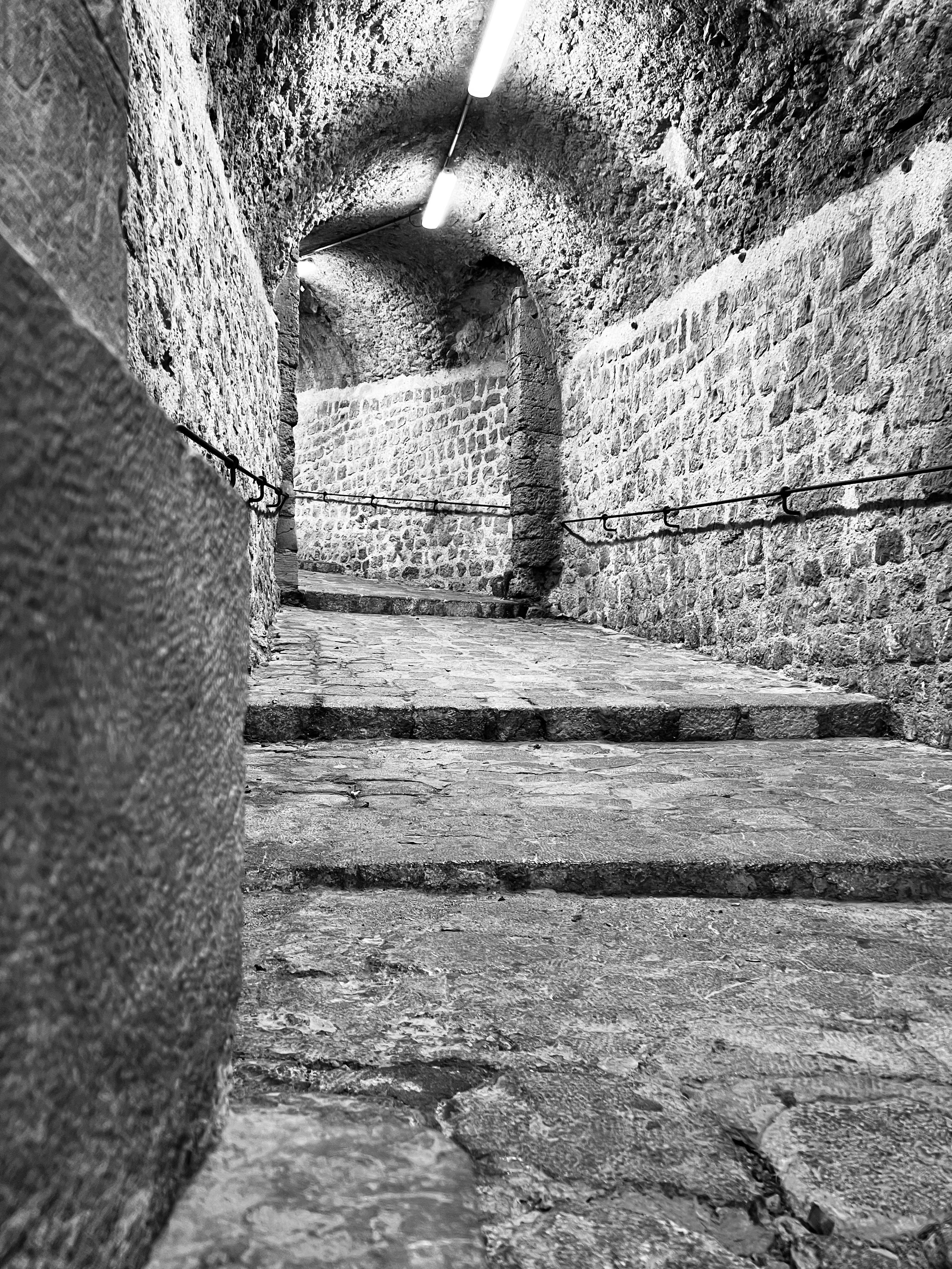 Black and white stone arched tunnel with illuminated steps and handrail.