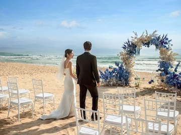 a man and woman standing on a beach with chairs and flowers