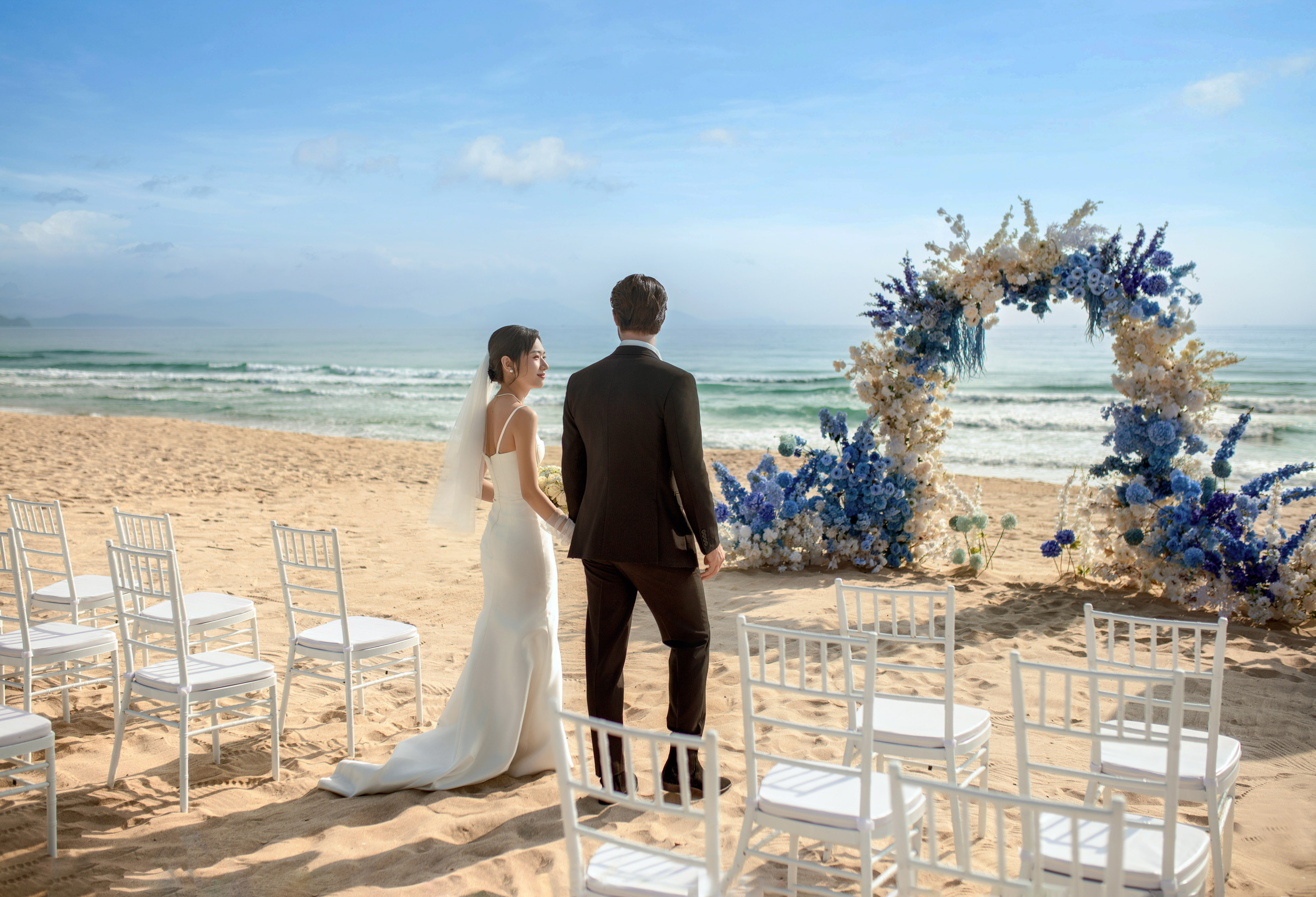 Bride and groom at beach wedding, facing ocean with blue floral arch.