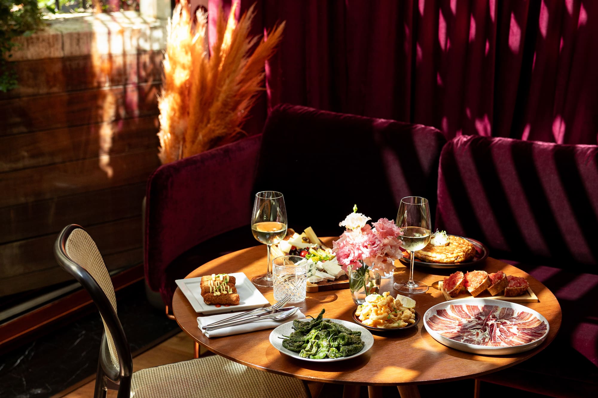 Restaurant table with assorted tapas, wine, and flowers in warm sunlight.