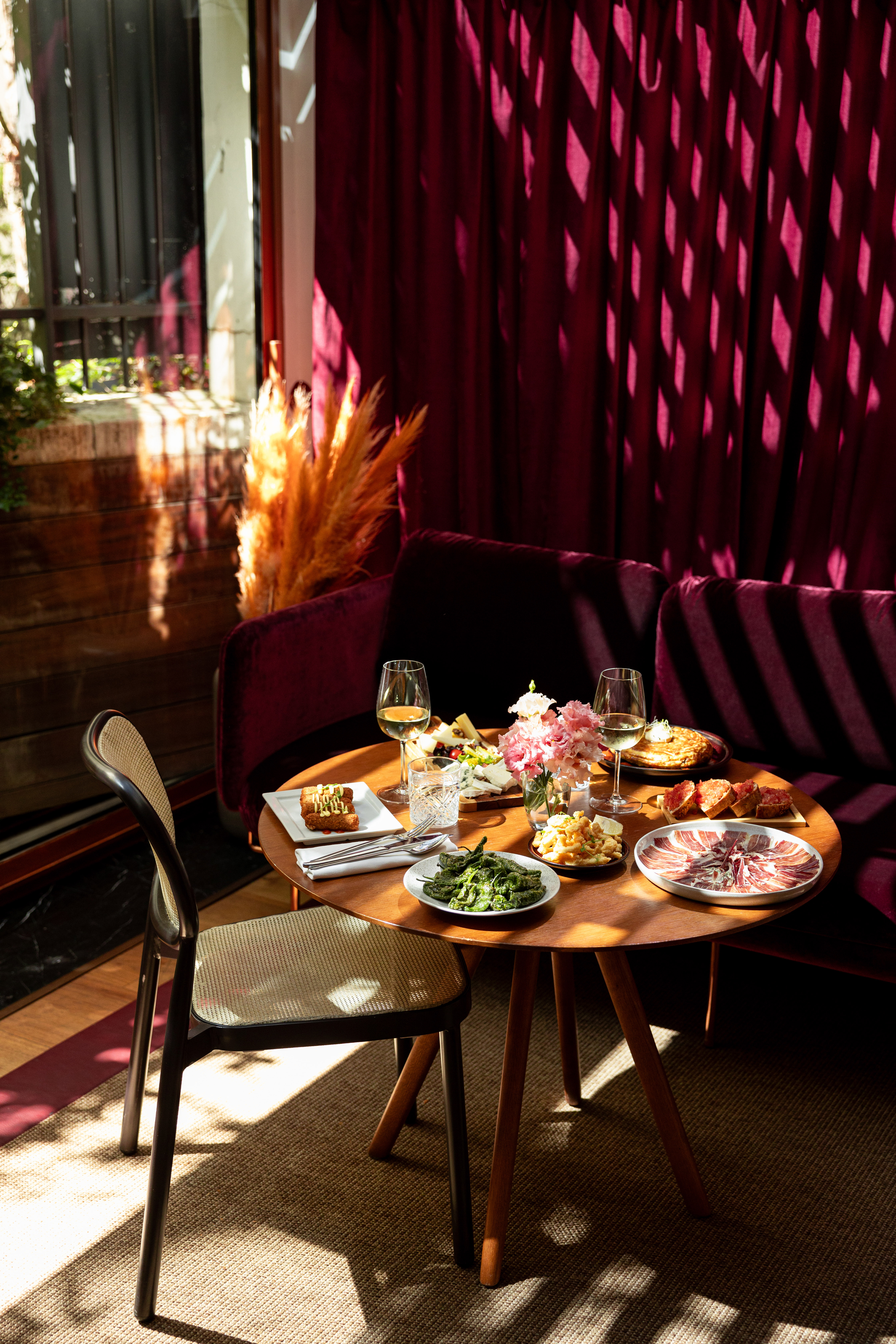 Restaurant table with assorted tapas, wine, and flowers in warm sunlight.