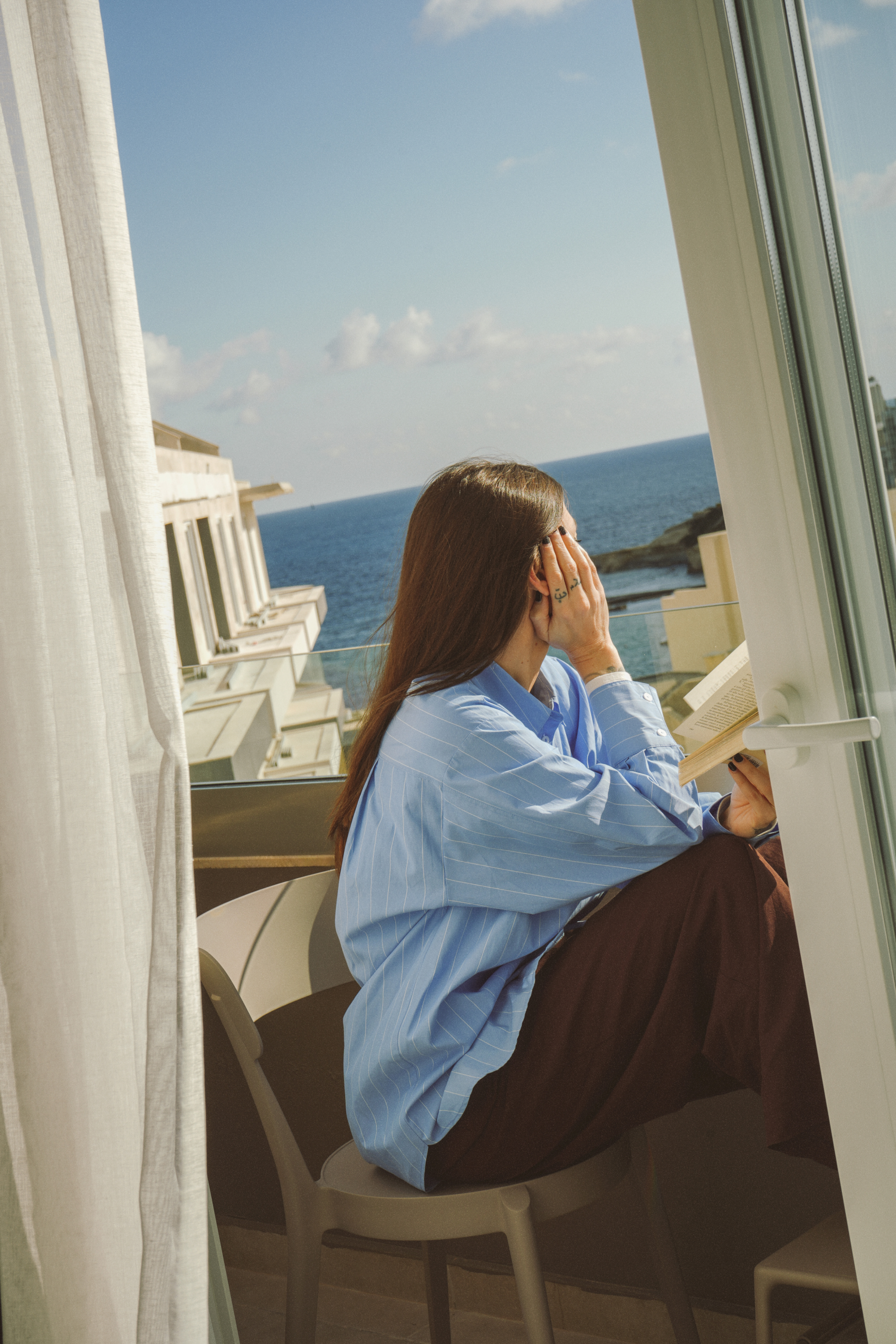 Woman in blue striped shirt reading book on resort balcony with ocean view.