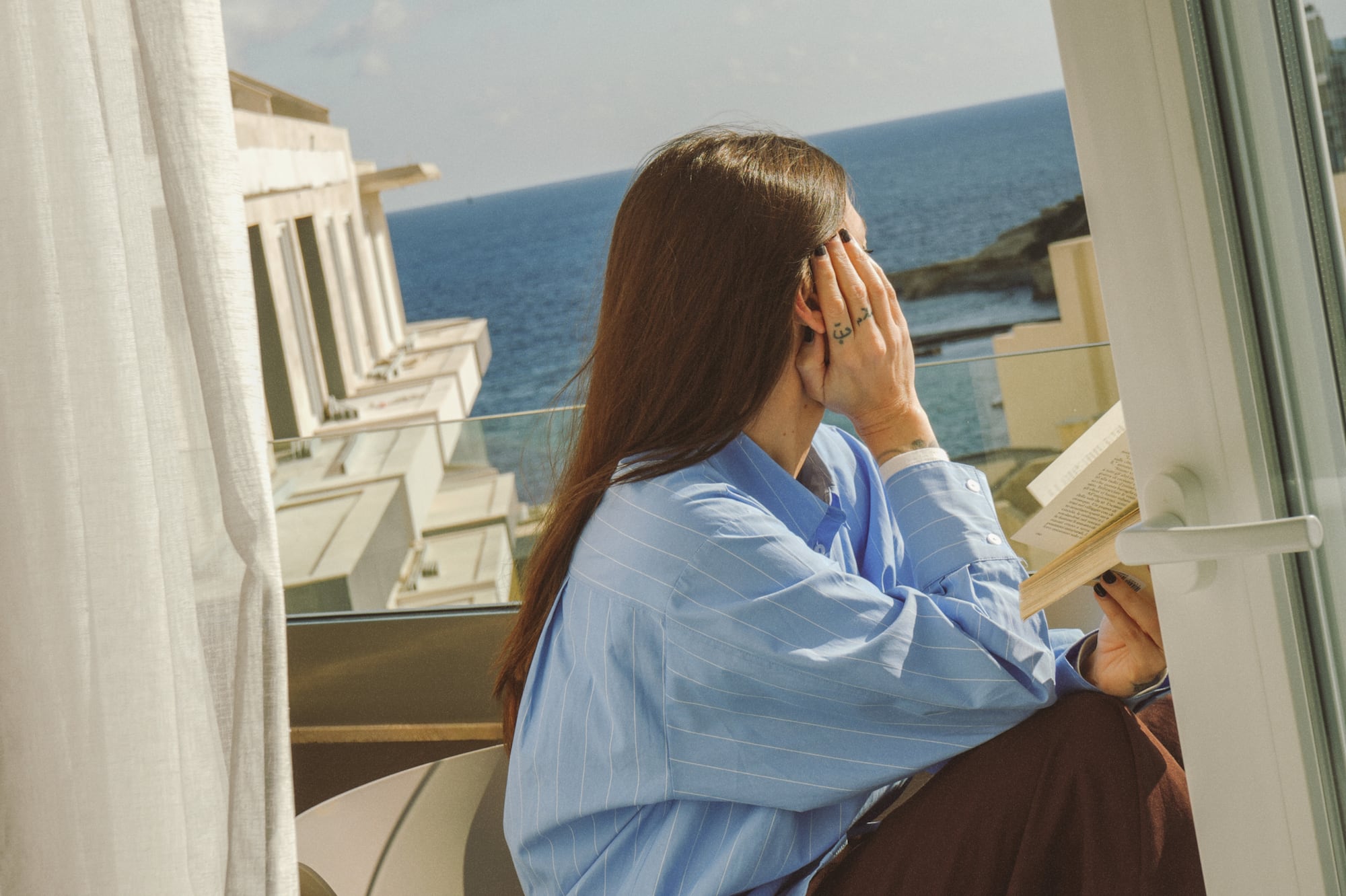 Woman in blue striped shirt reading book on resort balcony with ocean view.