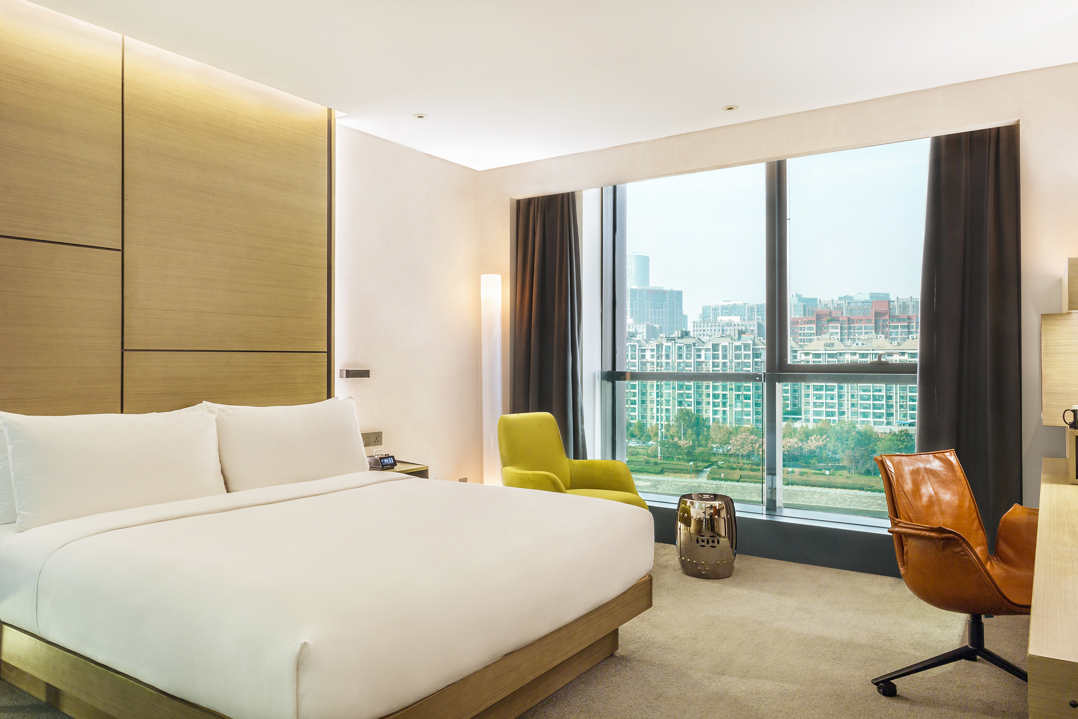 Contemporary hotel room, king bed, panoramic city view, lime armchair, workspace.