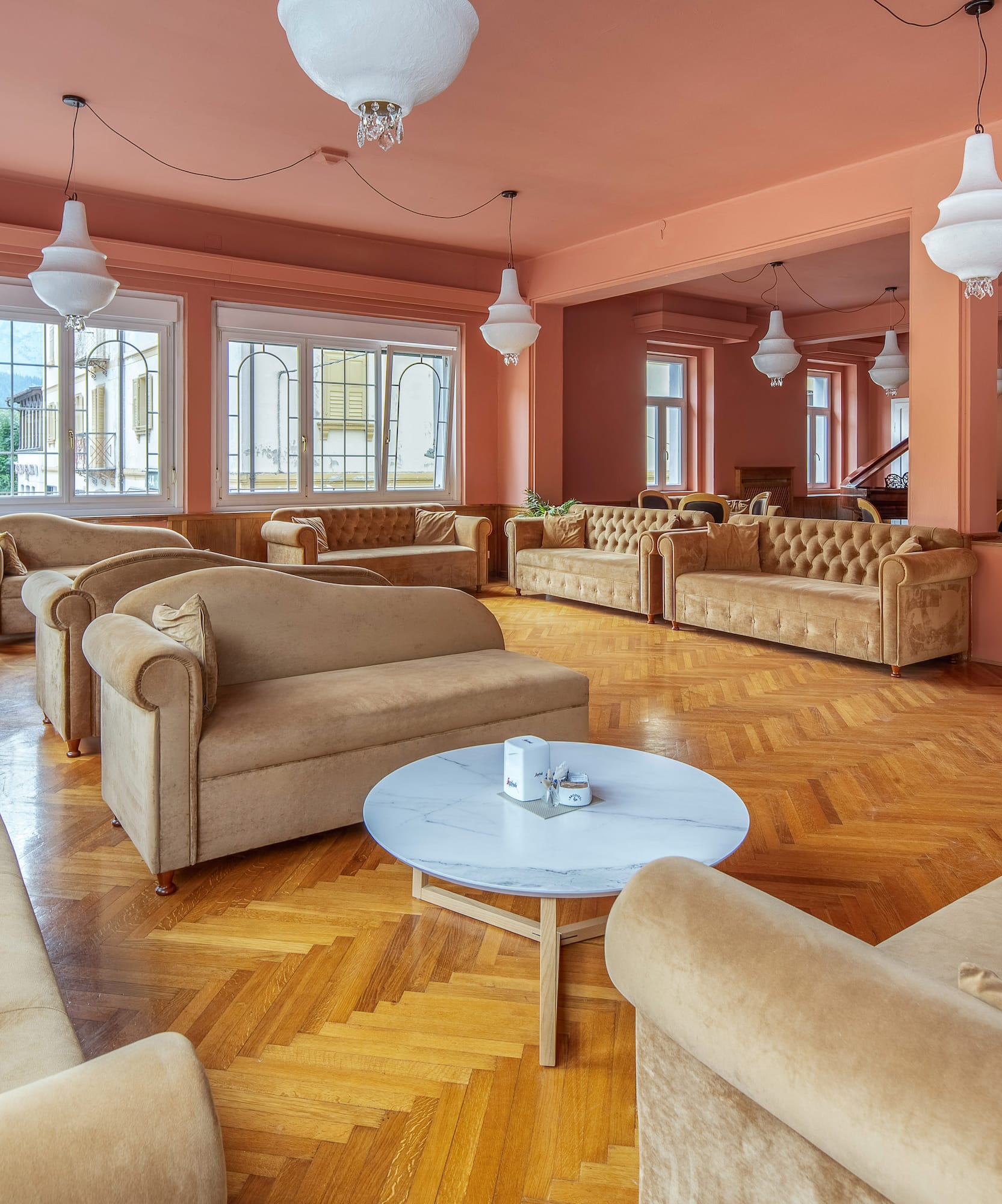 Peach hotel lounge, beige sofas, parquet floor, white lighting, large windows.
