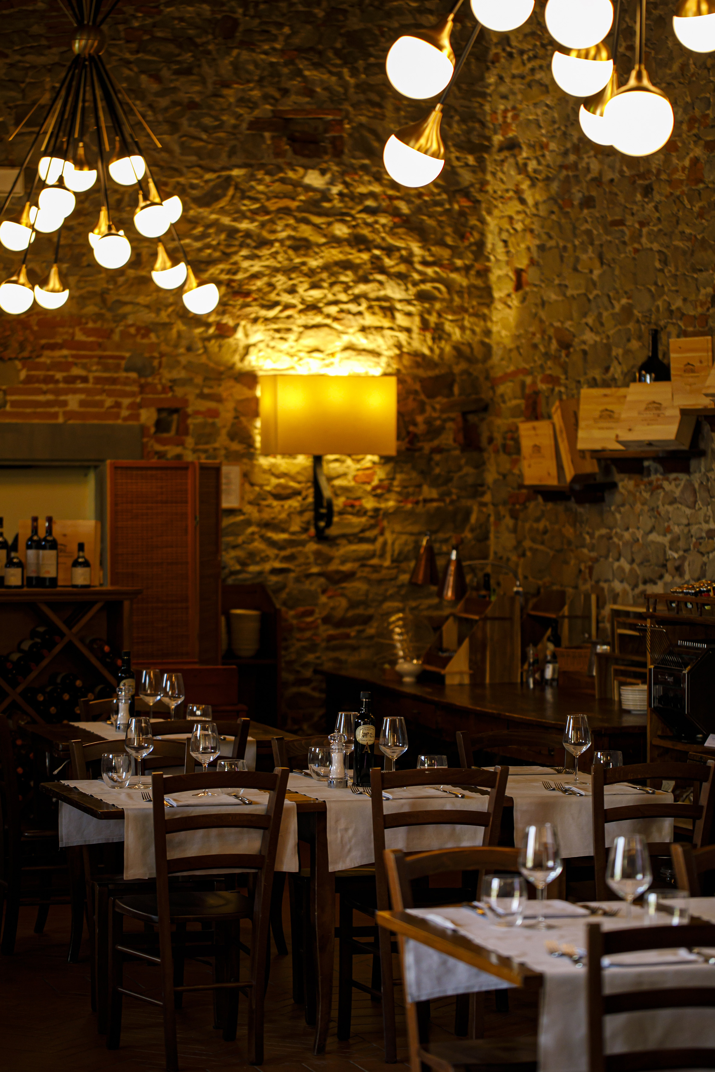 Cozy restaurant with stone walls, set tables, and warm, modern chandeliers.