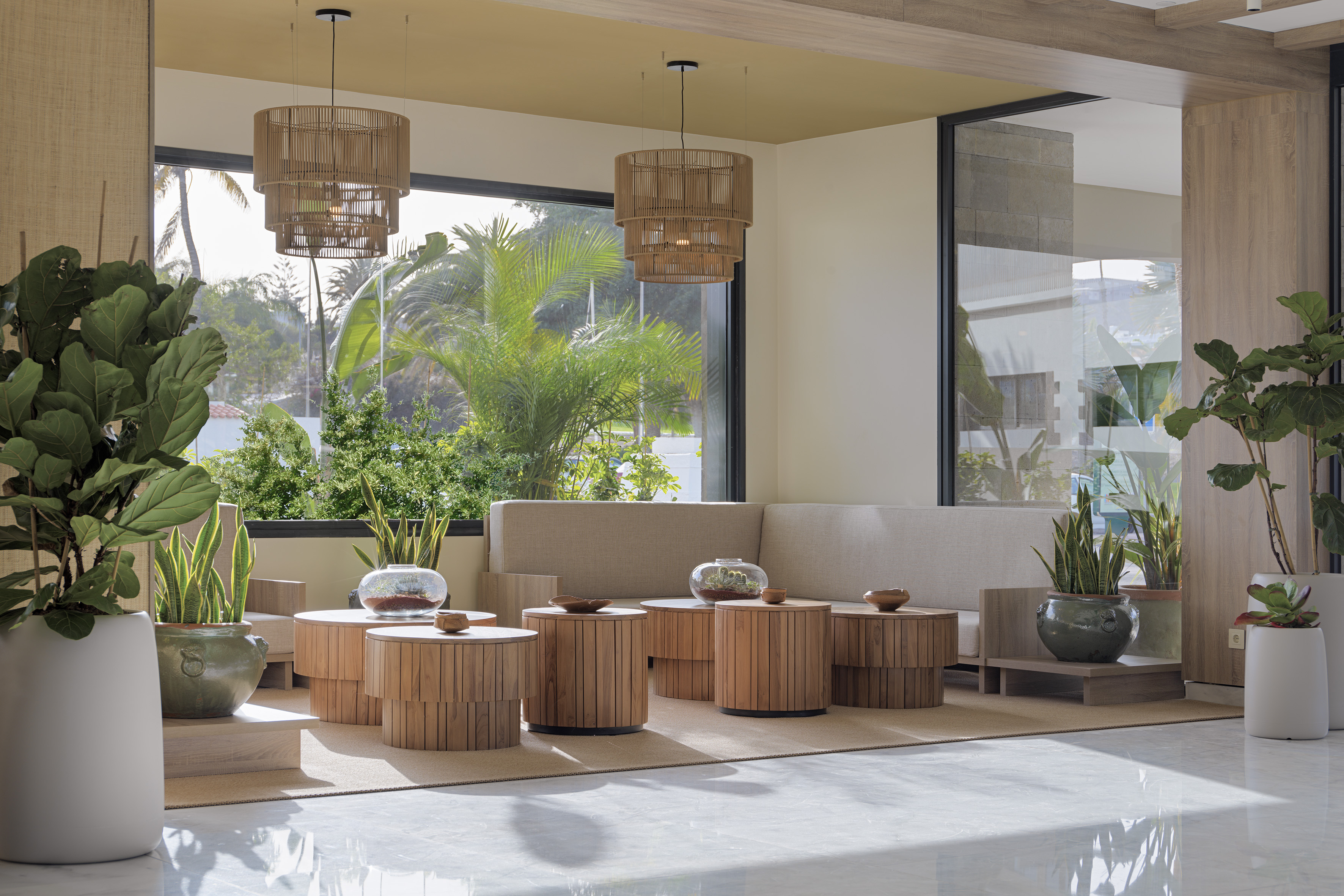 Modern hotel lounge with tropical plants, rattan lights, and wooden tables by large windows.