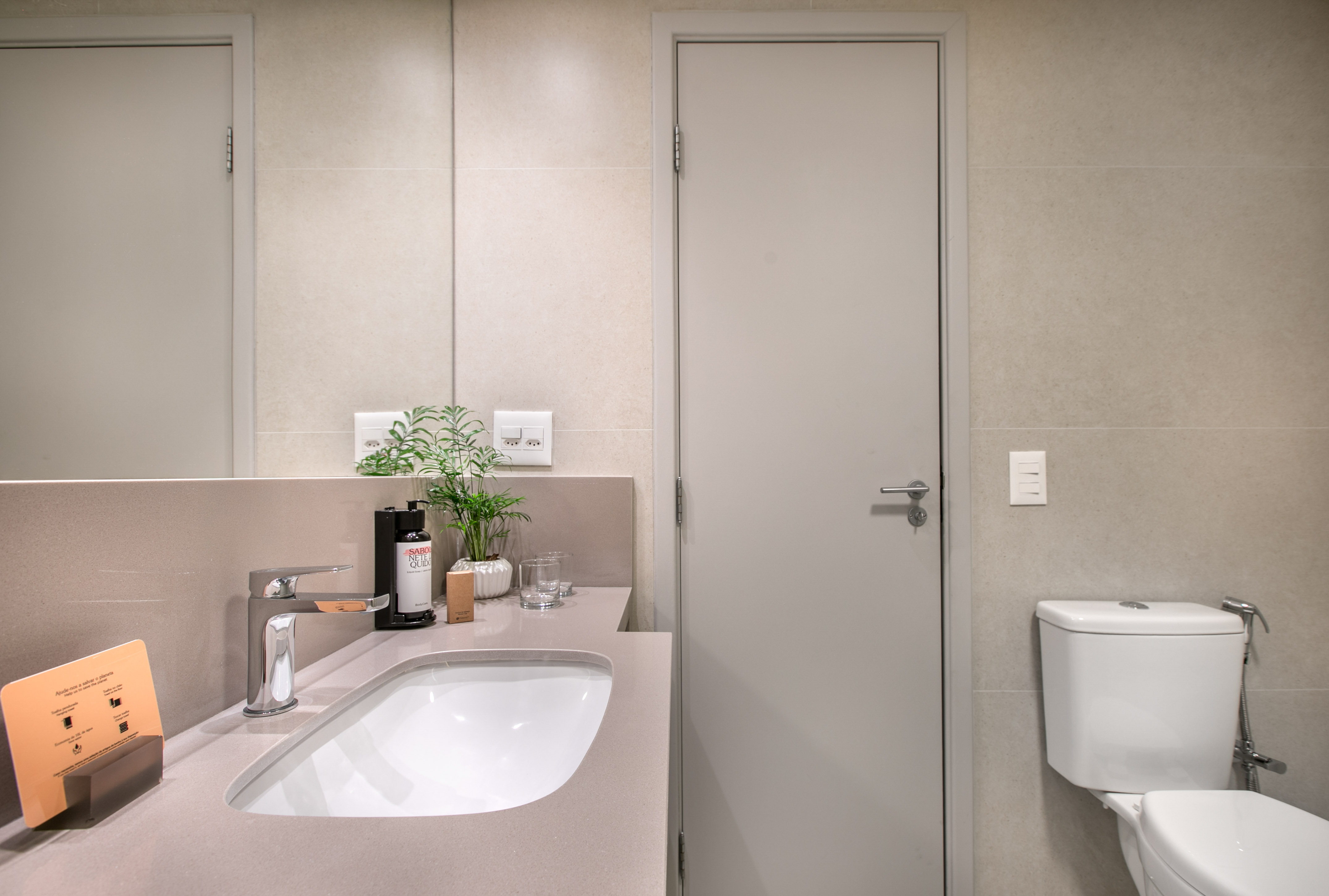 Modern hotel bathroom with gray vanity, sink, chrome faucet, plant, and toilet.