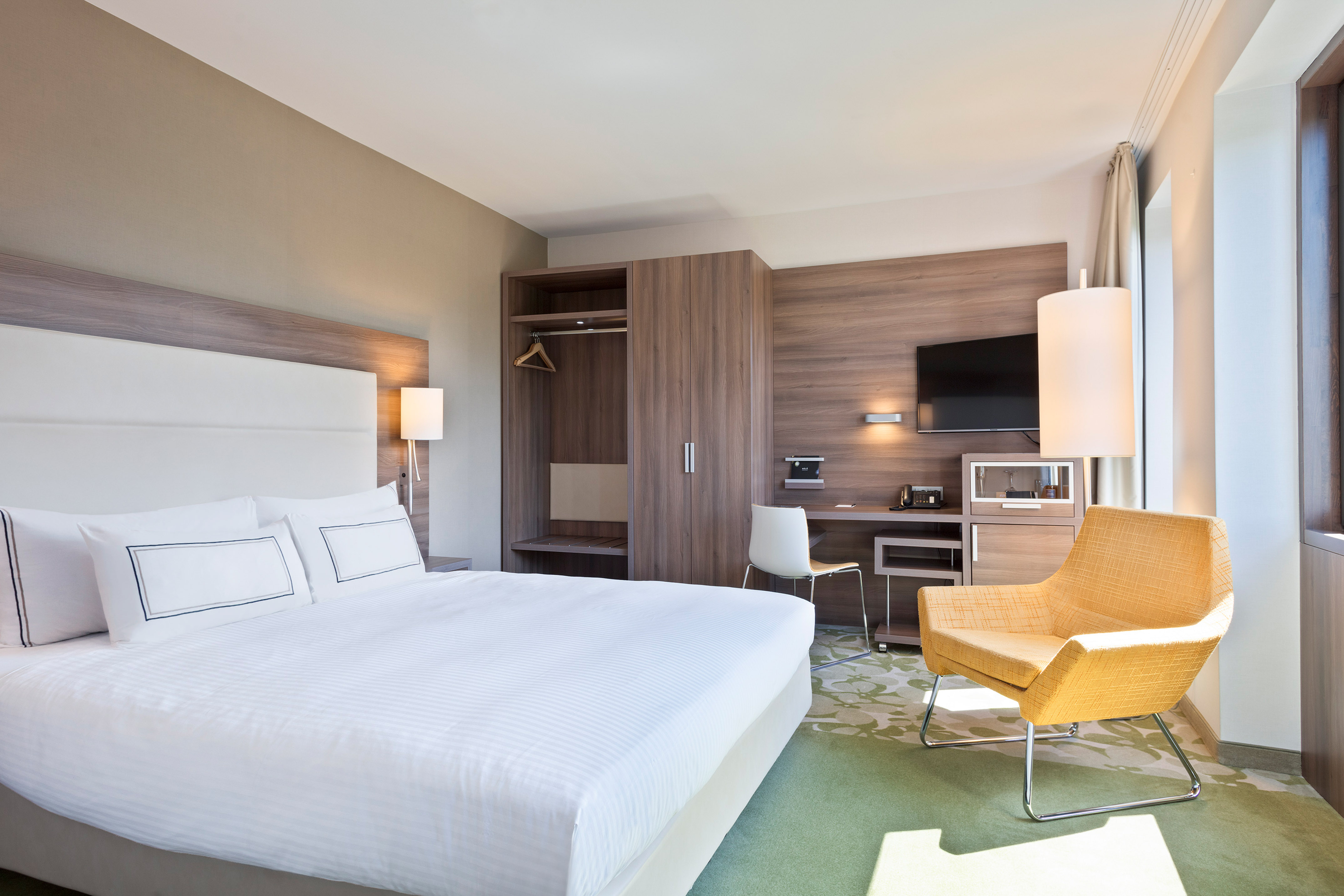 Modern hotel room with a large bed, wooden furniture, desk, and yellow armchair.