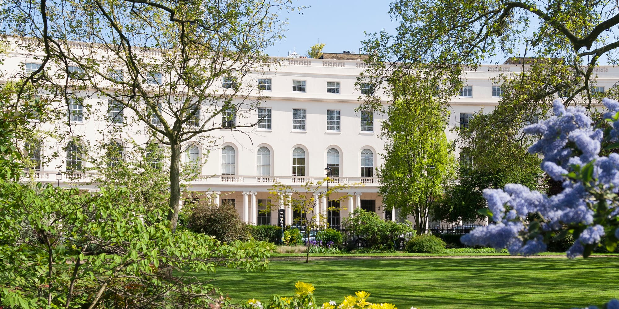 A white building featuring numerous windows stands next to a lawn dotted with trees under a clear blue sky.