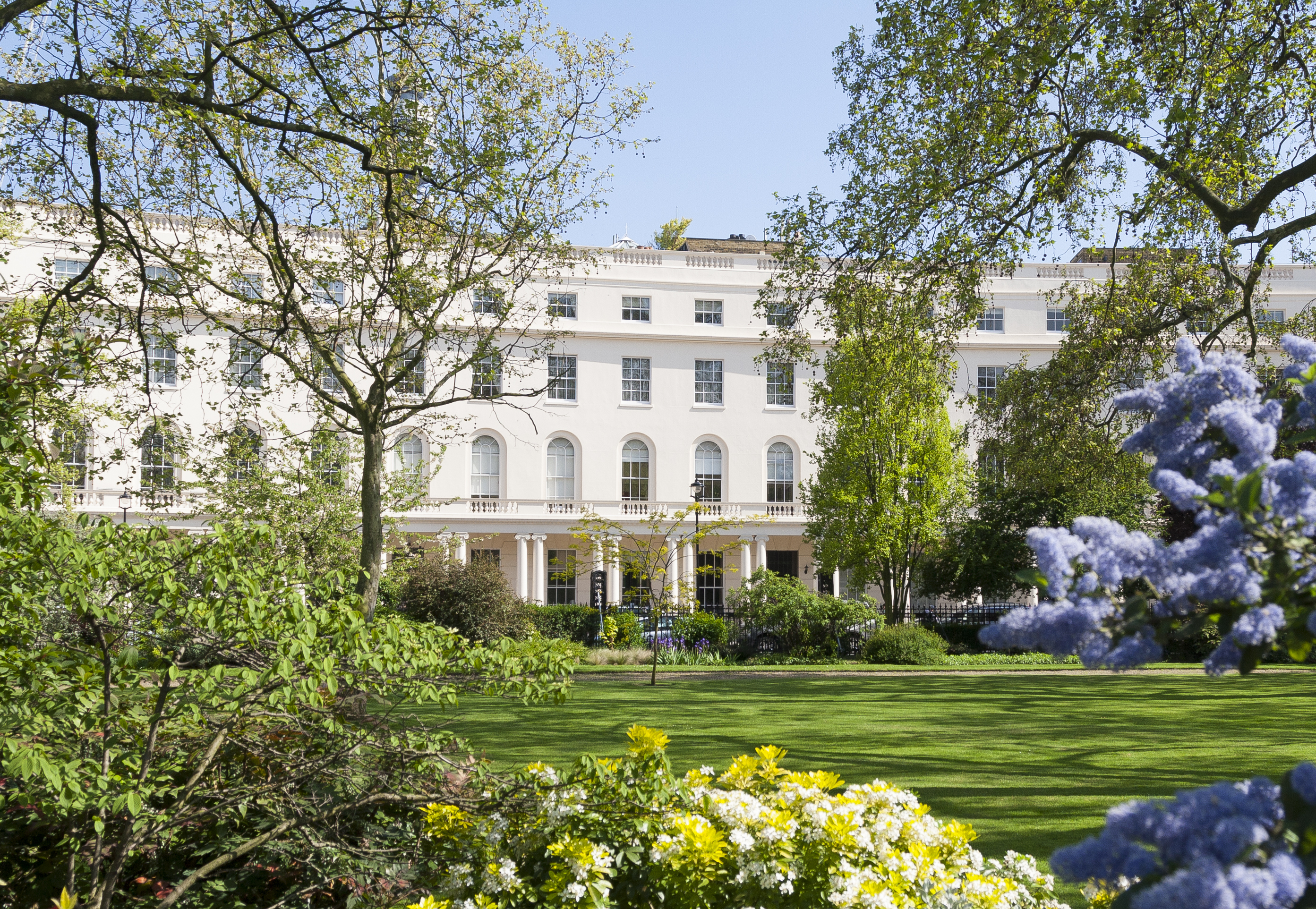 A white building featuring numerous windows, surrounded by a lawn dotted with trees under a clear blue sky.