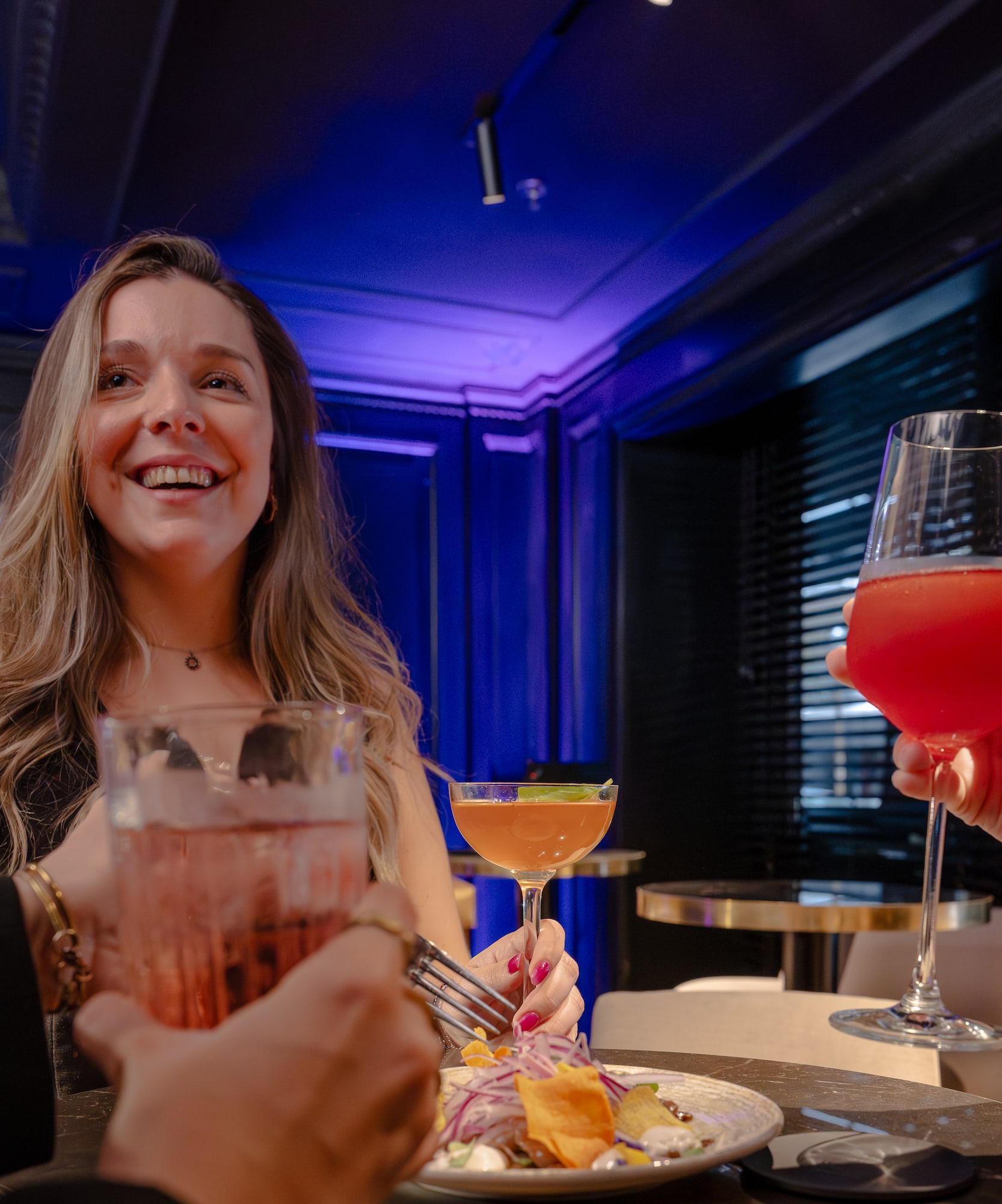 Mujer sonriente y amigos disfrutando cócteles y platillos en un bar elegante.