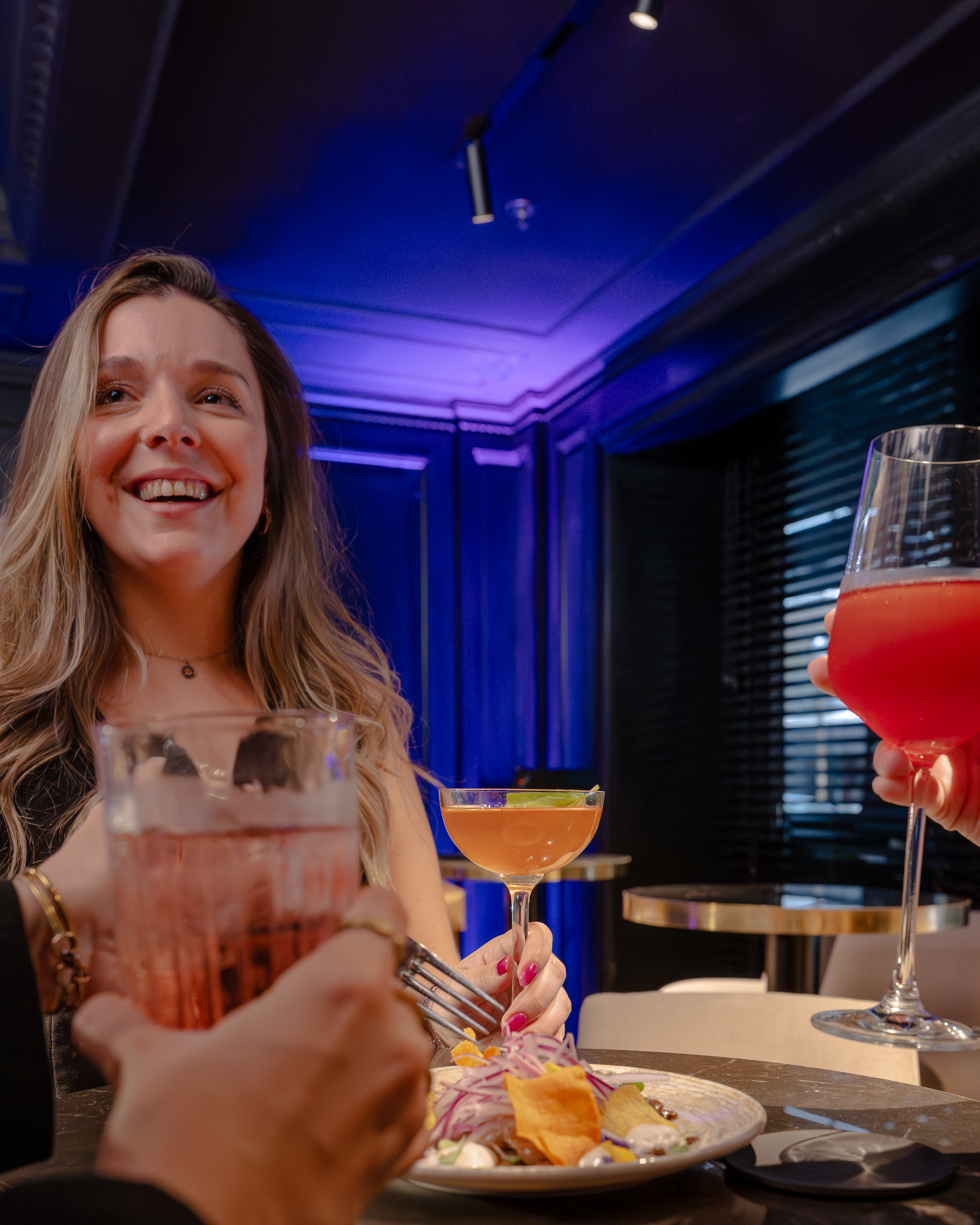 Mujer sonriente y amigos disfrutando cócteles y platillos en un bar elegante.