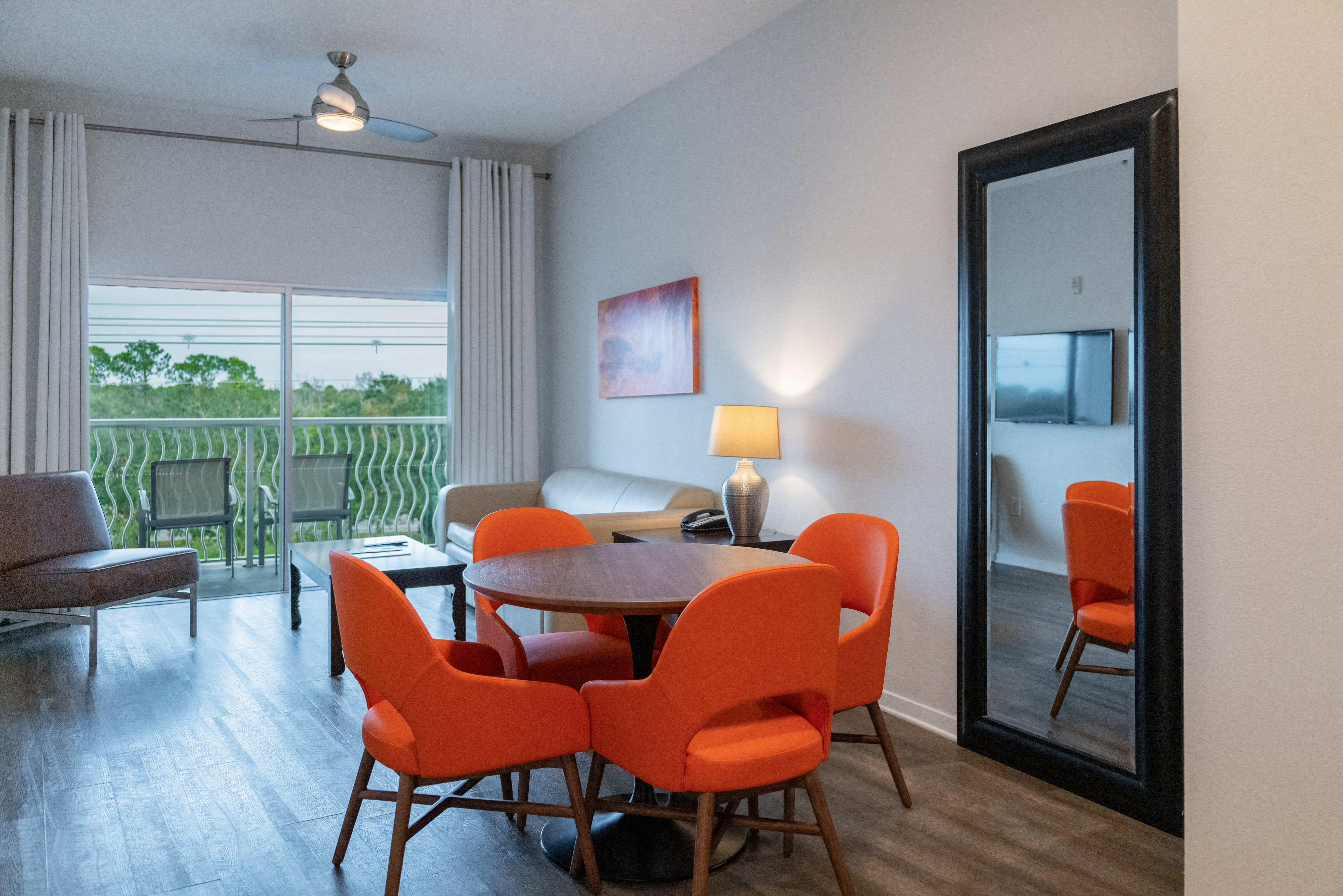 Modern living and dining room with orange chairs, sofa, balcony, and wood floors.