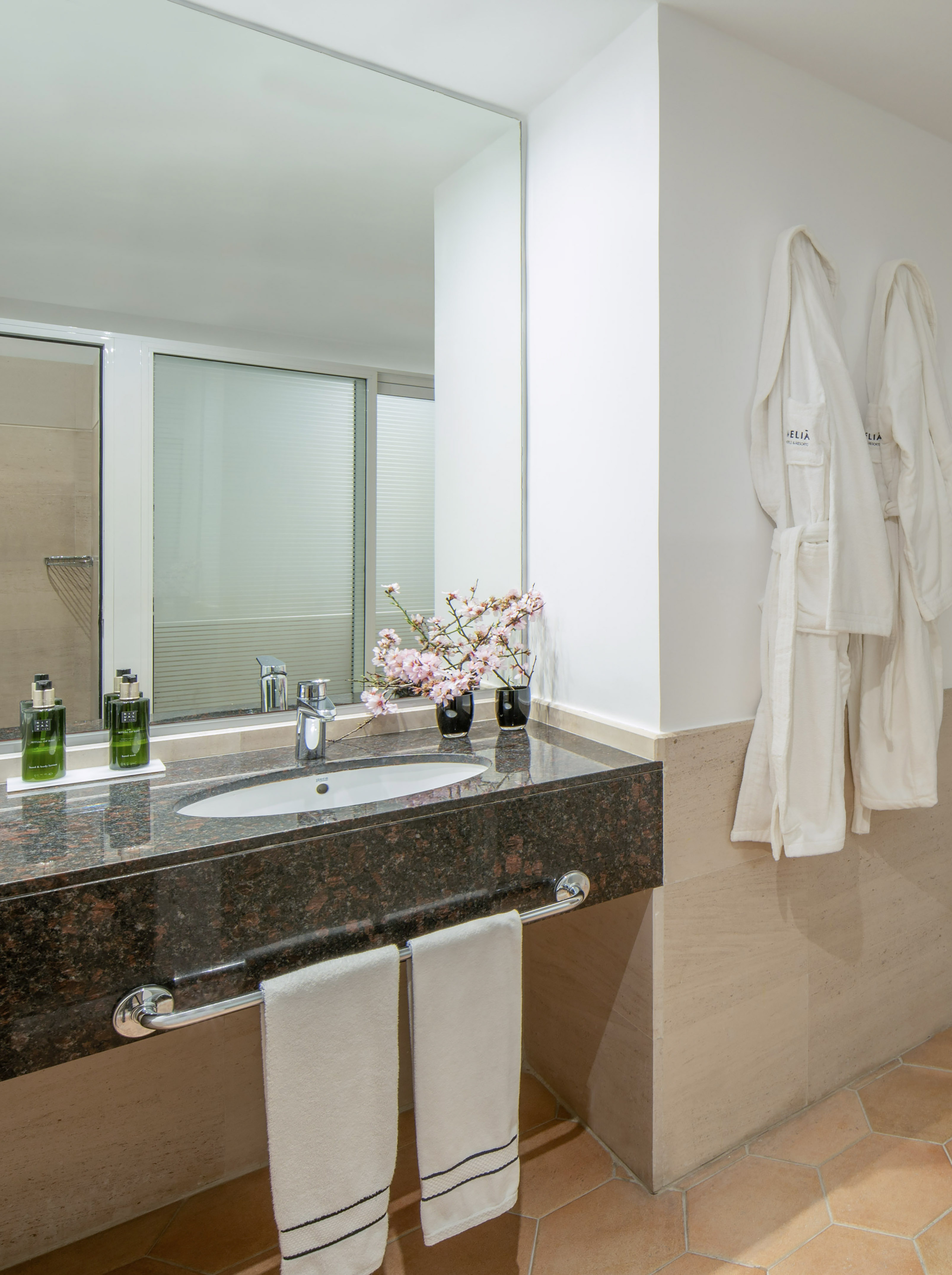 Hotel bathroom with granite vanity, sink, mirror, toiletries, and two white bathrobes.