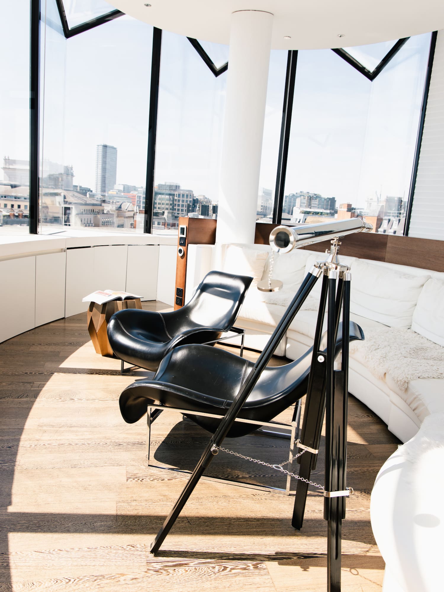 Modern living room, city view through panoramic windows, black chairs, and telescope.