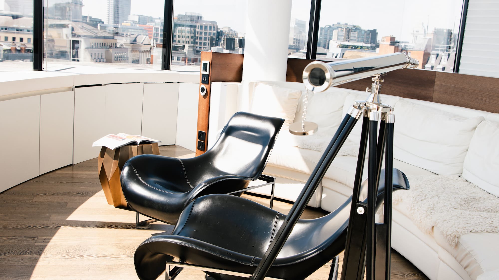 Bright modern living room with city views, black chairs, white sofa, and telescope.