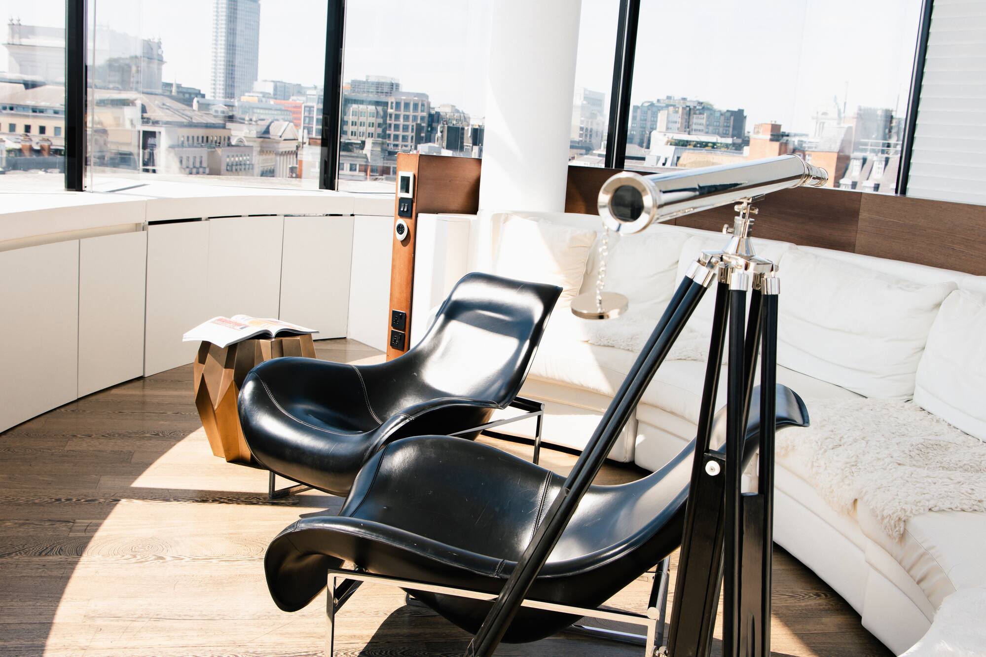 Bright modern living room with city views, black chairs, white sofa, and telescope.