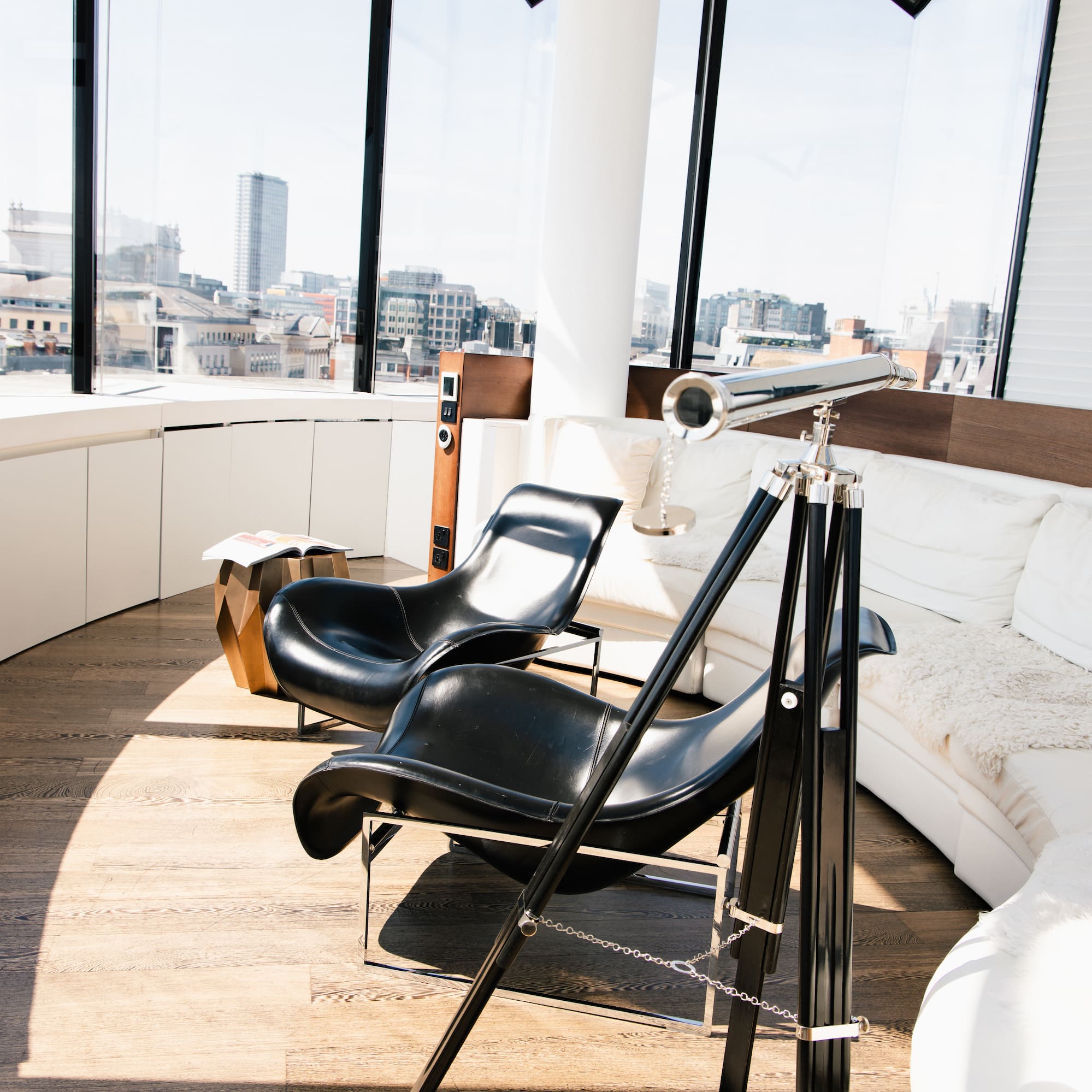 Bright modern living room with city views, black chairs, white sofa, and telescope.