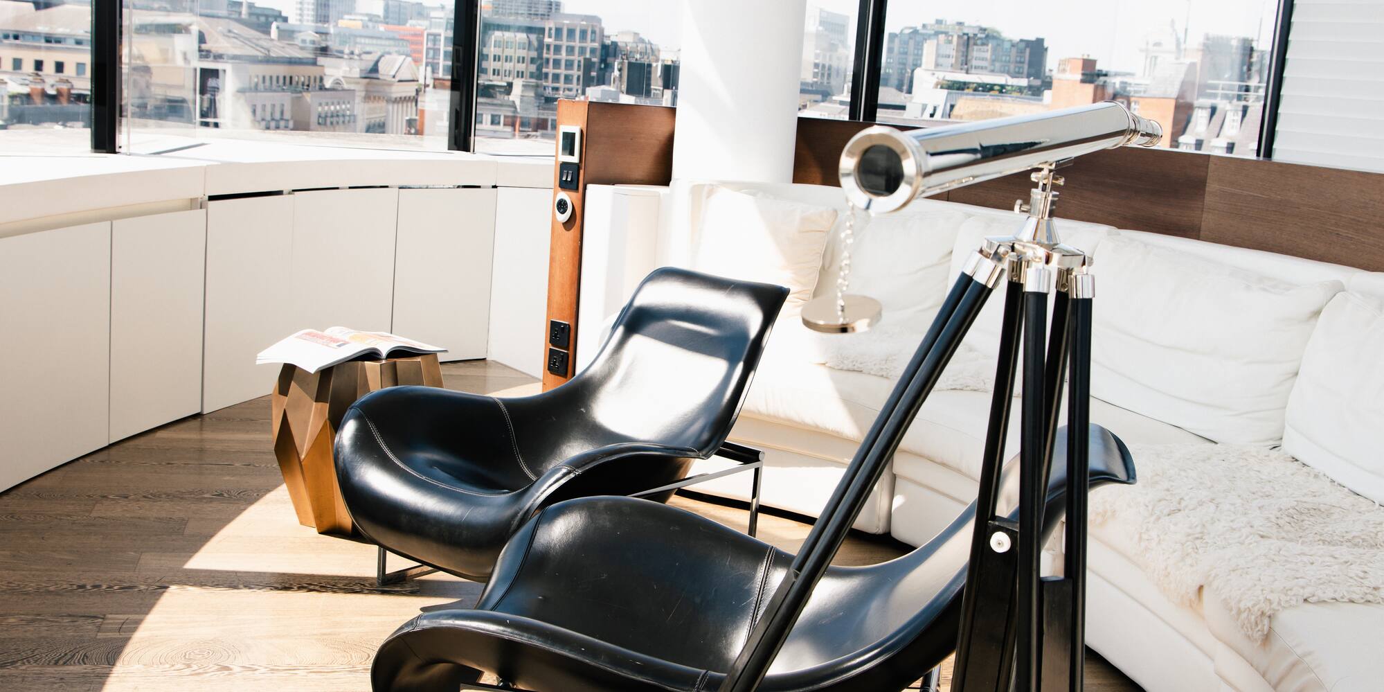 Bright modern living room with city views, black chairs, white sofa, and telescope.