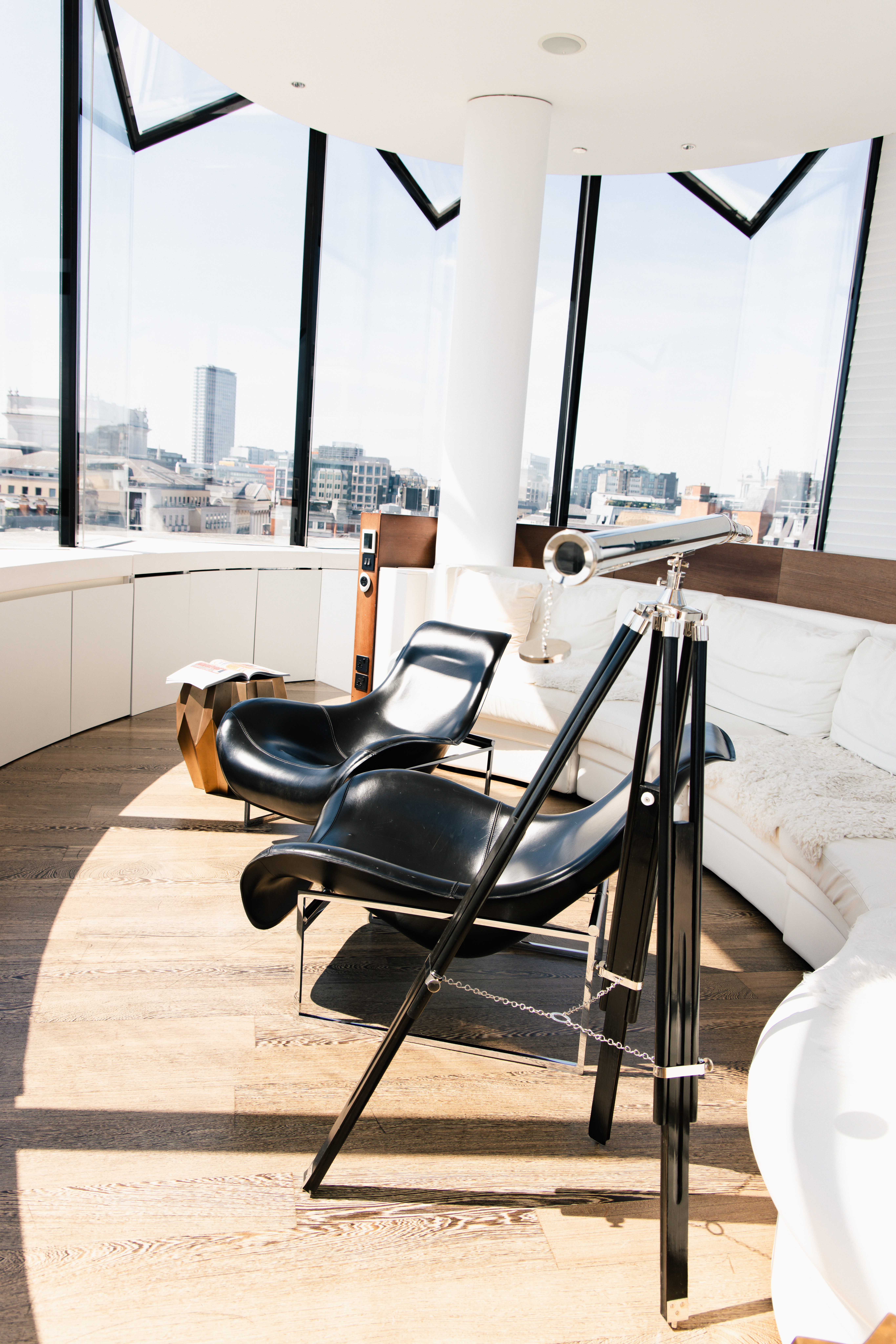 Bright modern living room with city views, black chairs, white sofa, and telescope.