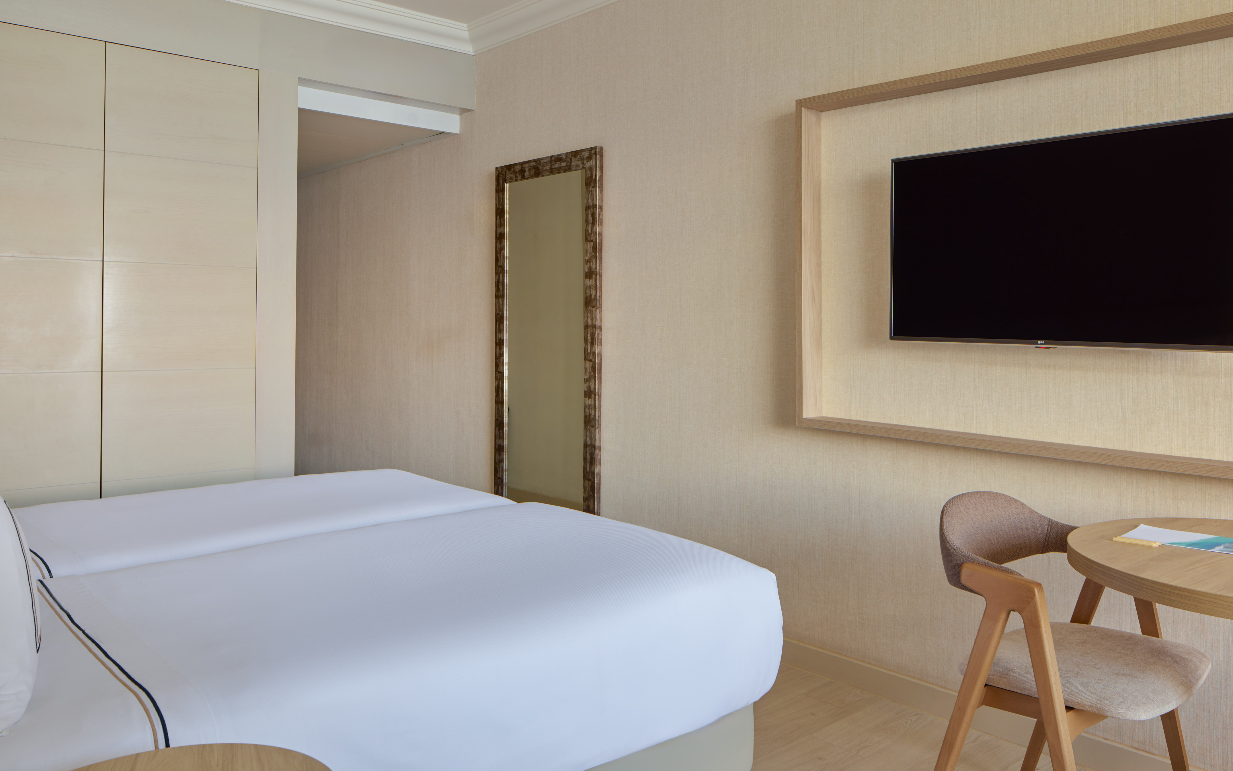 Hotel room with two beds, wall-mounted TV, wooden table, and full-length mirror.