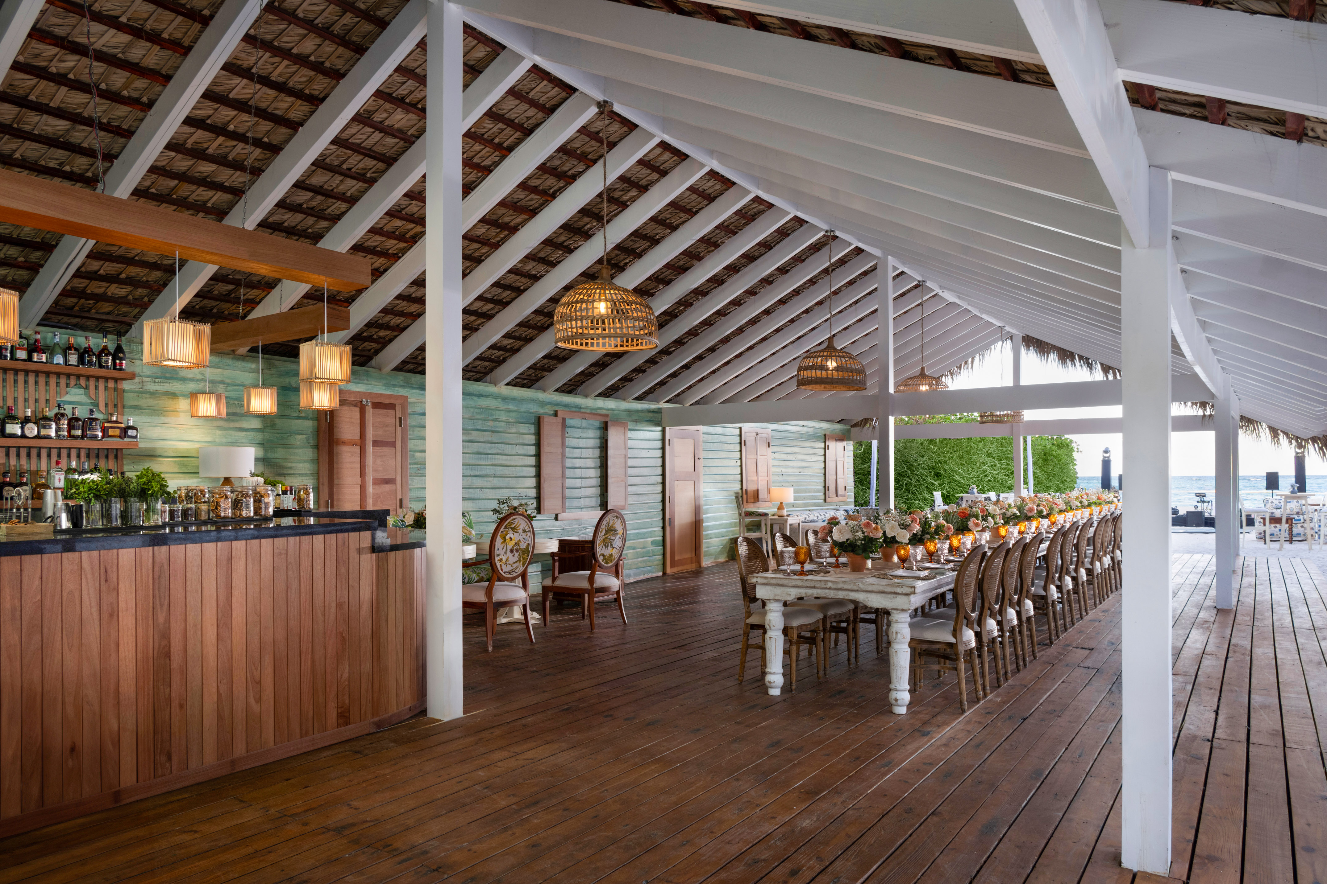 Tropical beach restaurant with long dining table, bar, and ocean views.