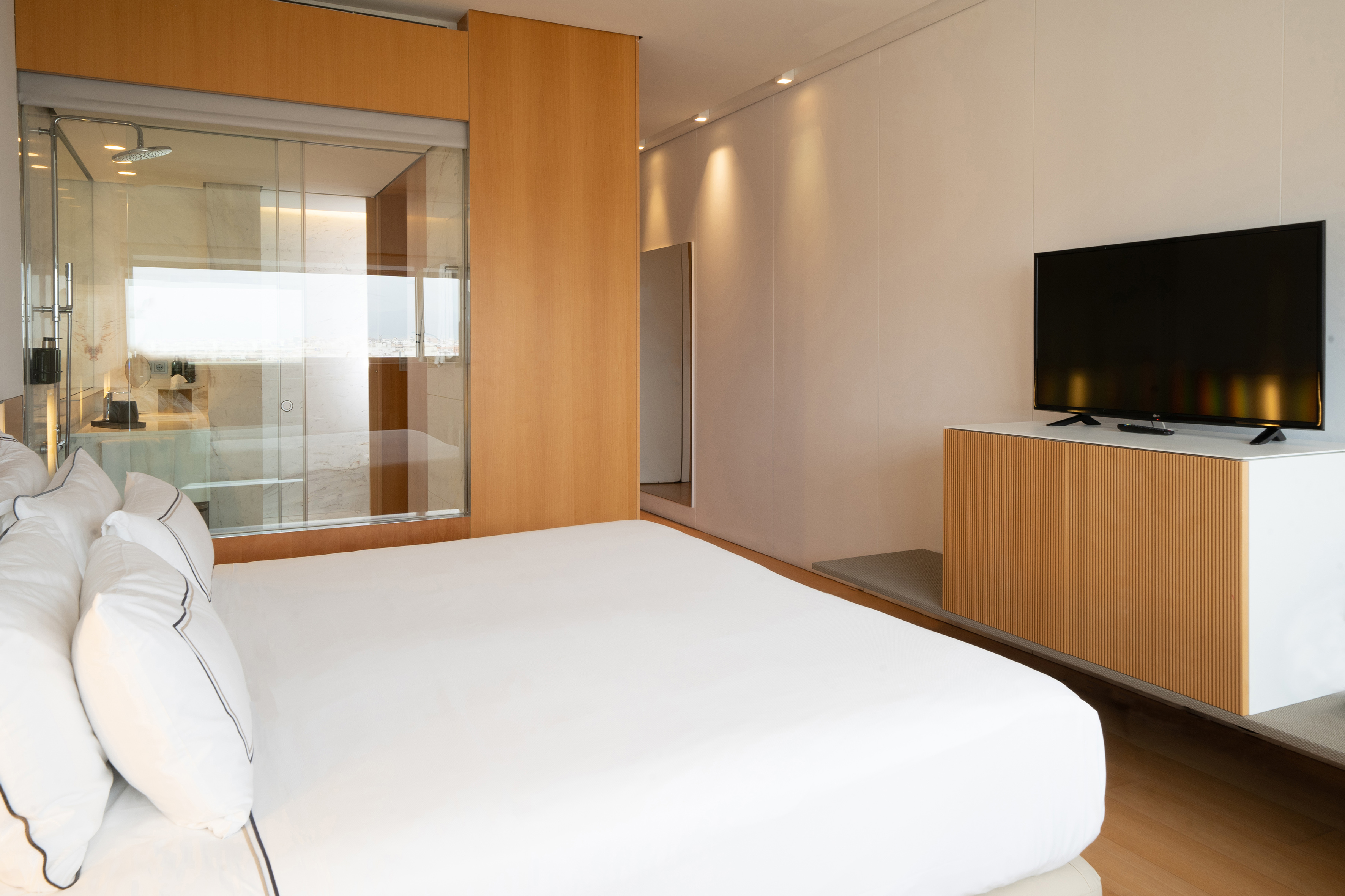 Modern hotel suite with white bed, glass shower, wood accents, and flat-screen TV.