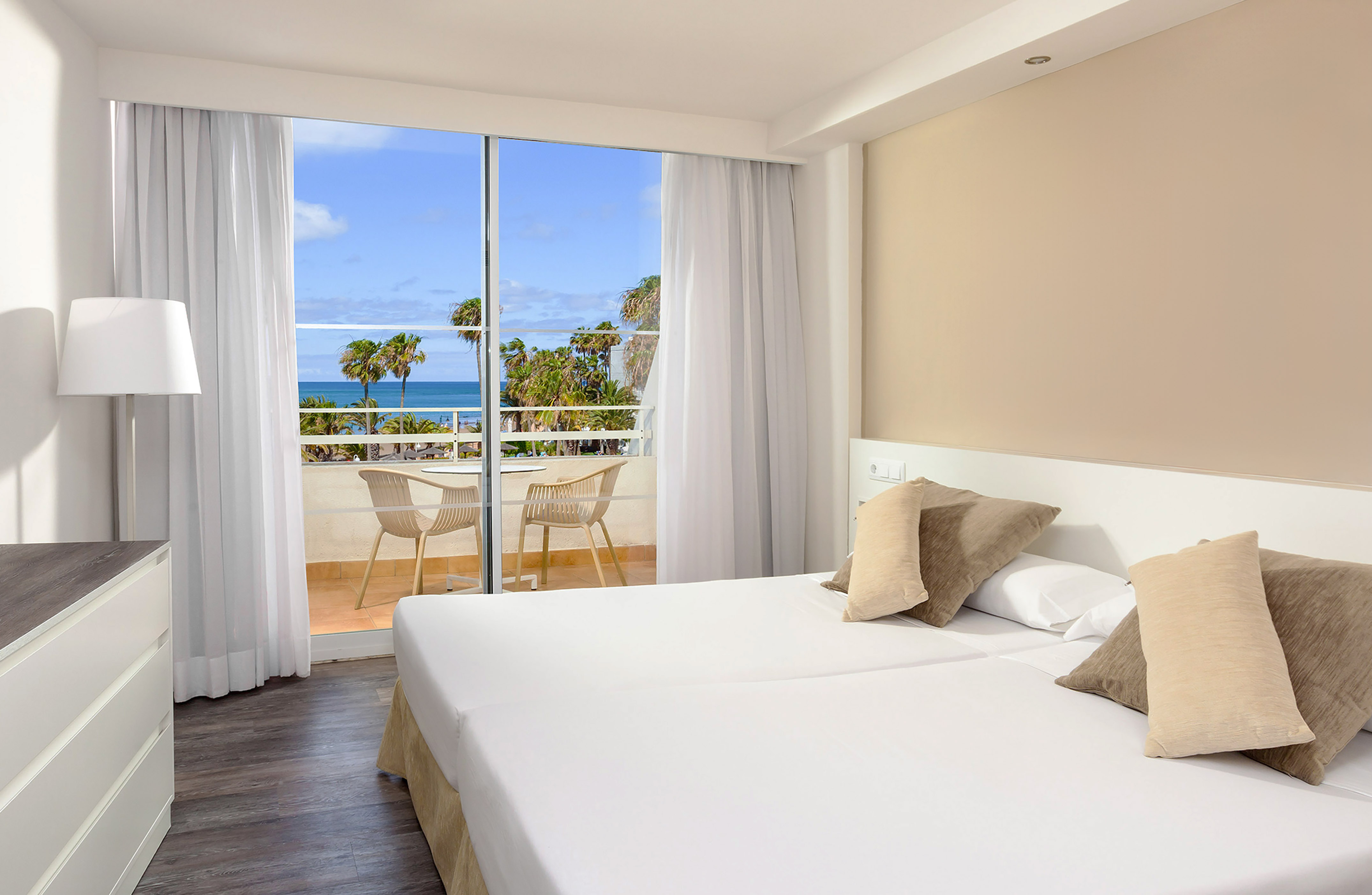 Hotel bedroom with ocean view, balcony, and tropical palm trees.