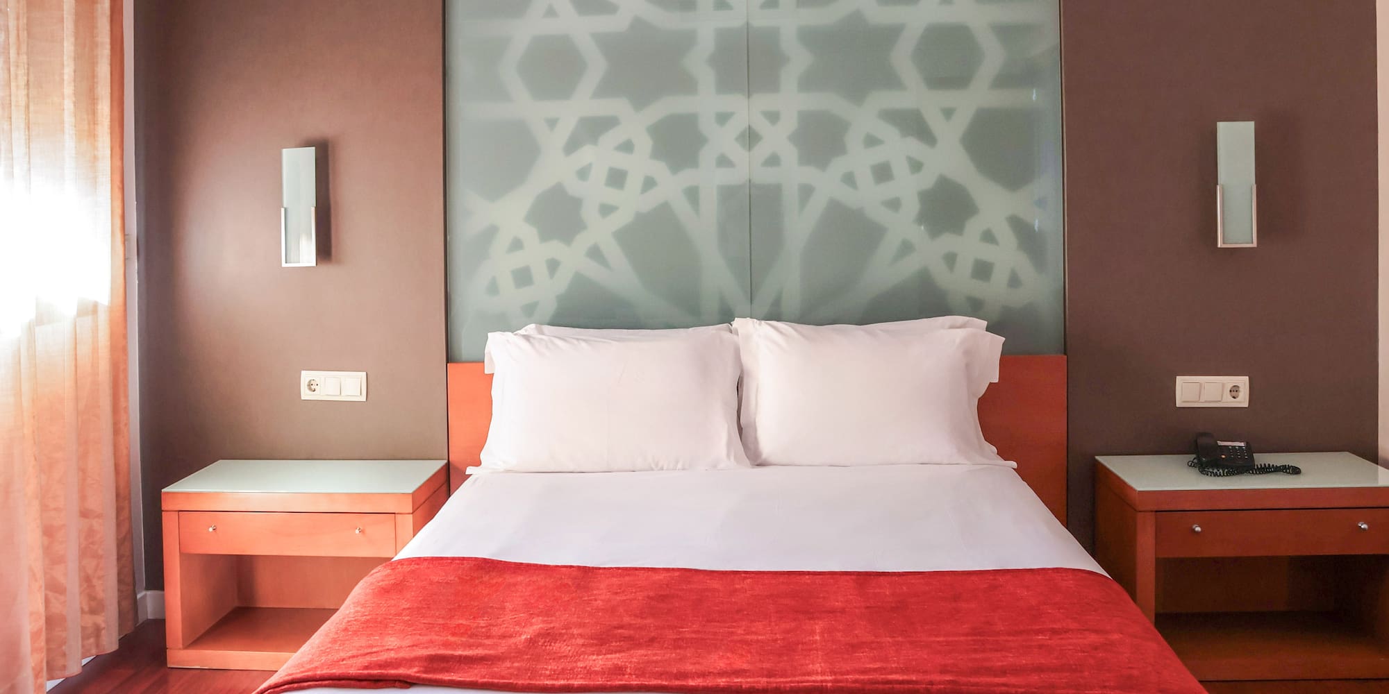 Hotel bedroom with white queen bed, red runner, and geometric glass headboard.