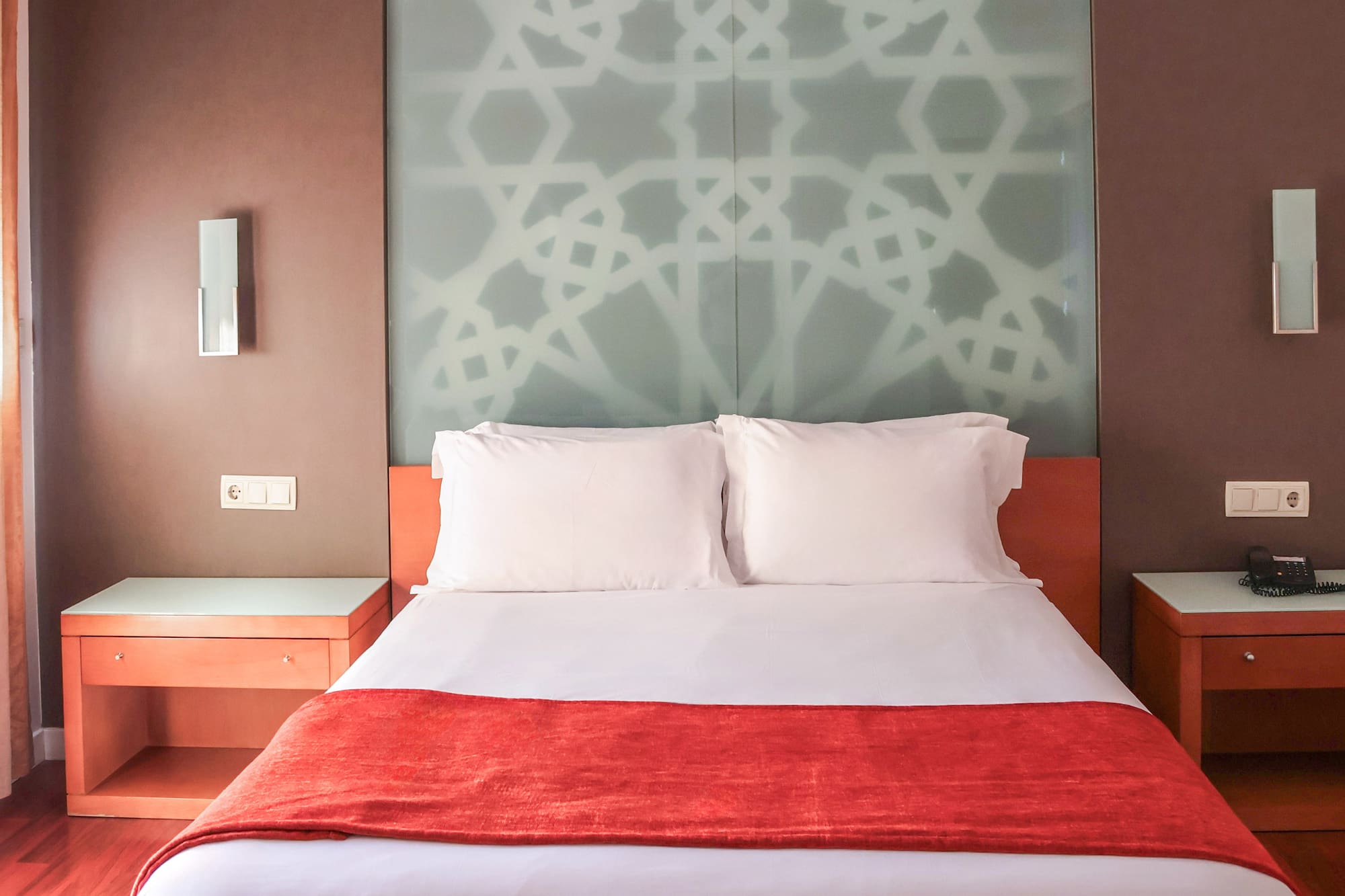 Hotel bedroom with white queen bed, red runner, and geometric glass headboard.