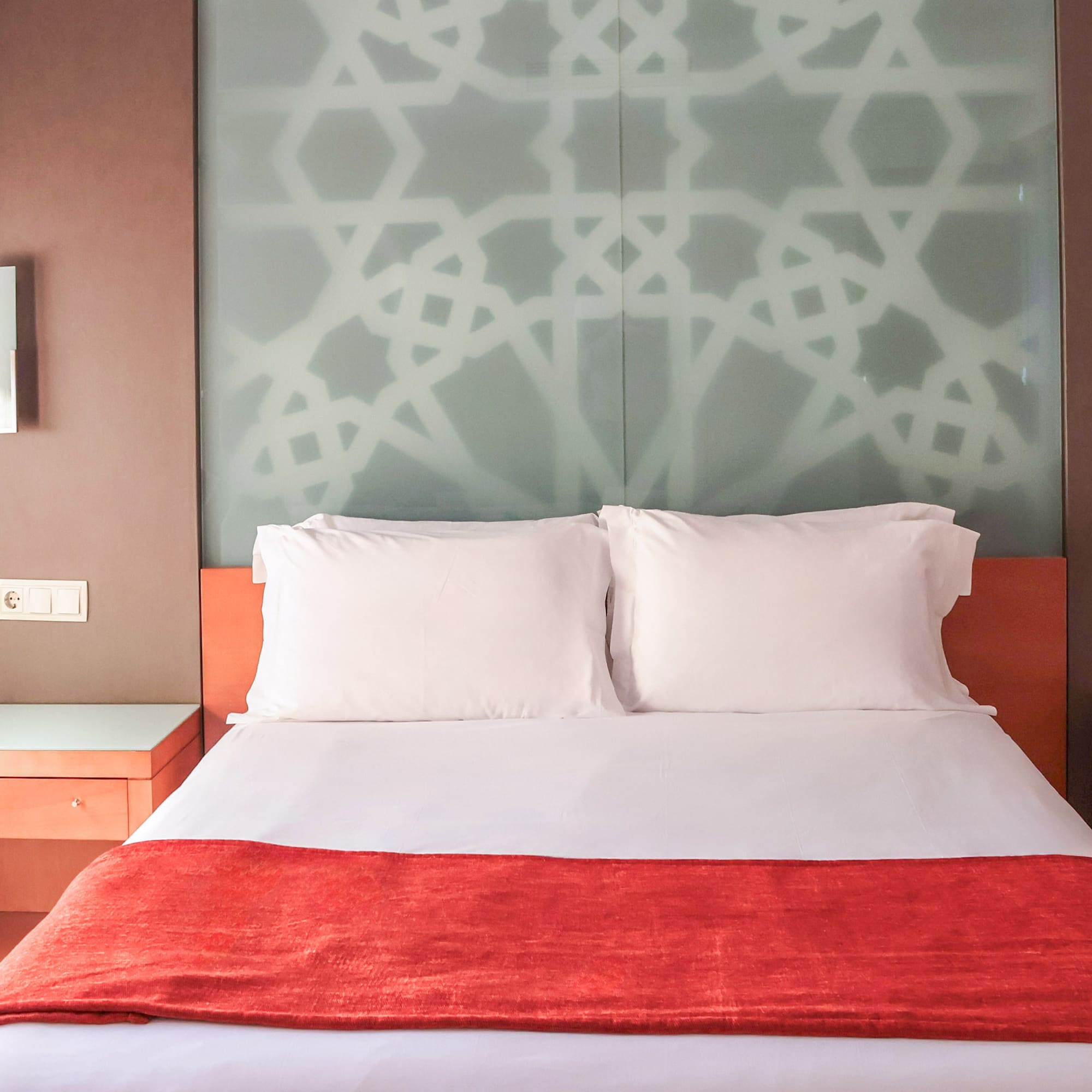 Hotel bedroom with white queen bed, red runner, and geometric glass headboard.