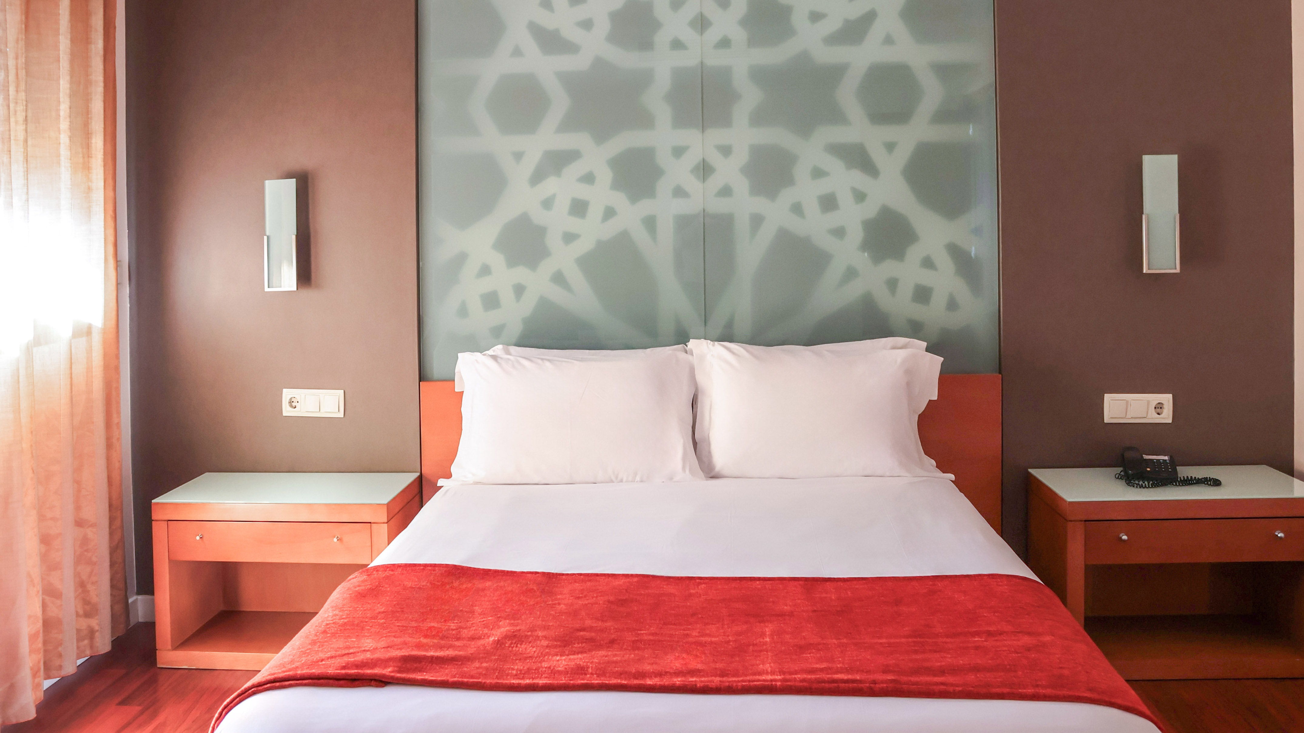 Hotel bedroom with white queen bed, red runner, and geometric glass headboard.
