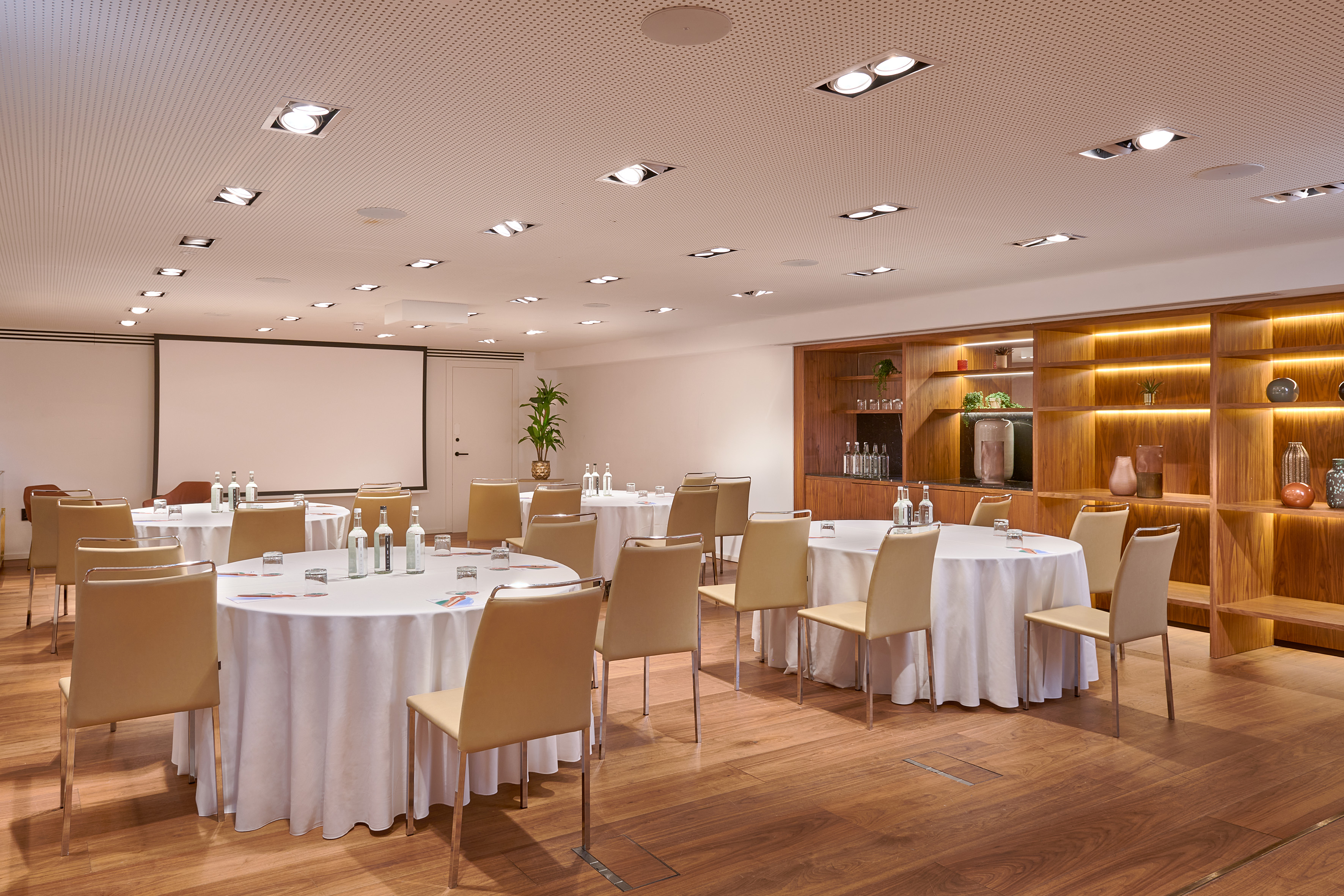 Hotel conference room with round tables, screen, and wood shelves.