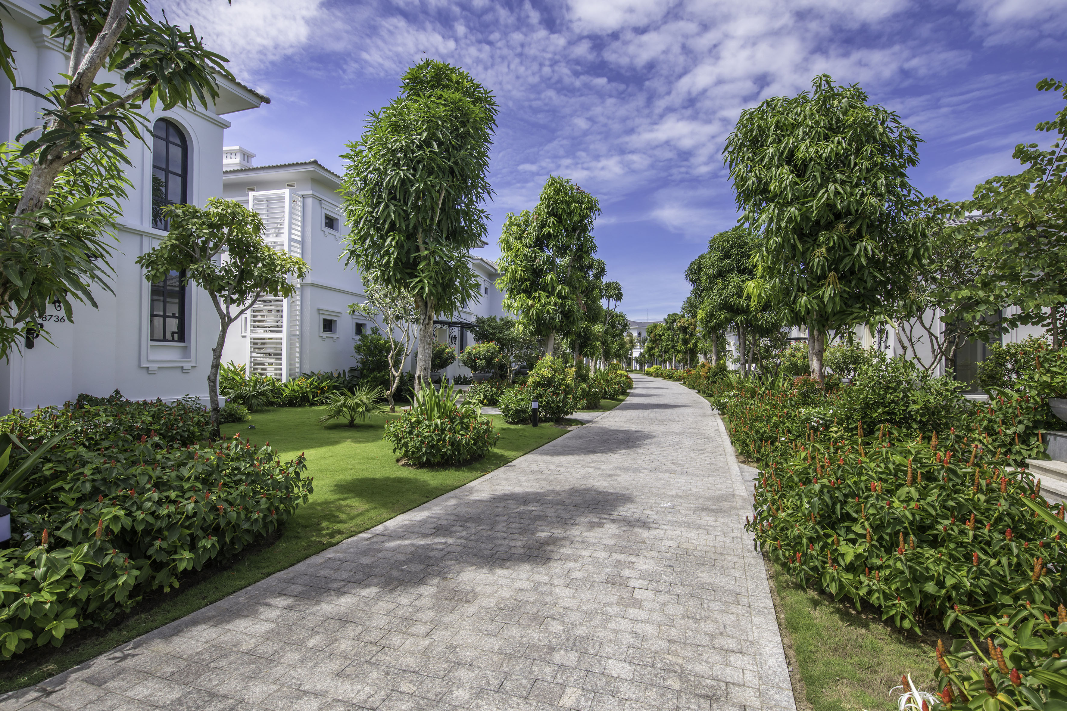 Modern white villas line a paved path with green lawns and tropical trees.